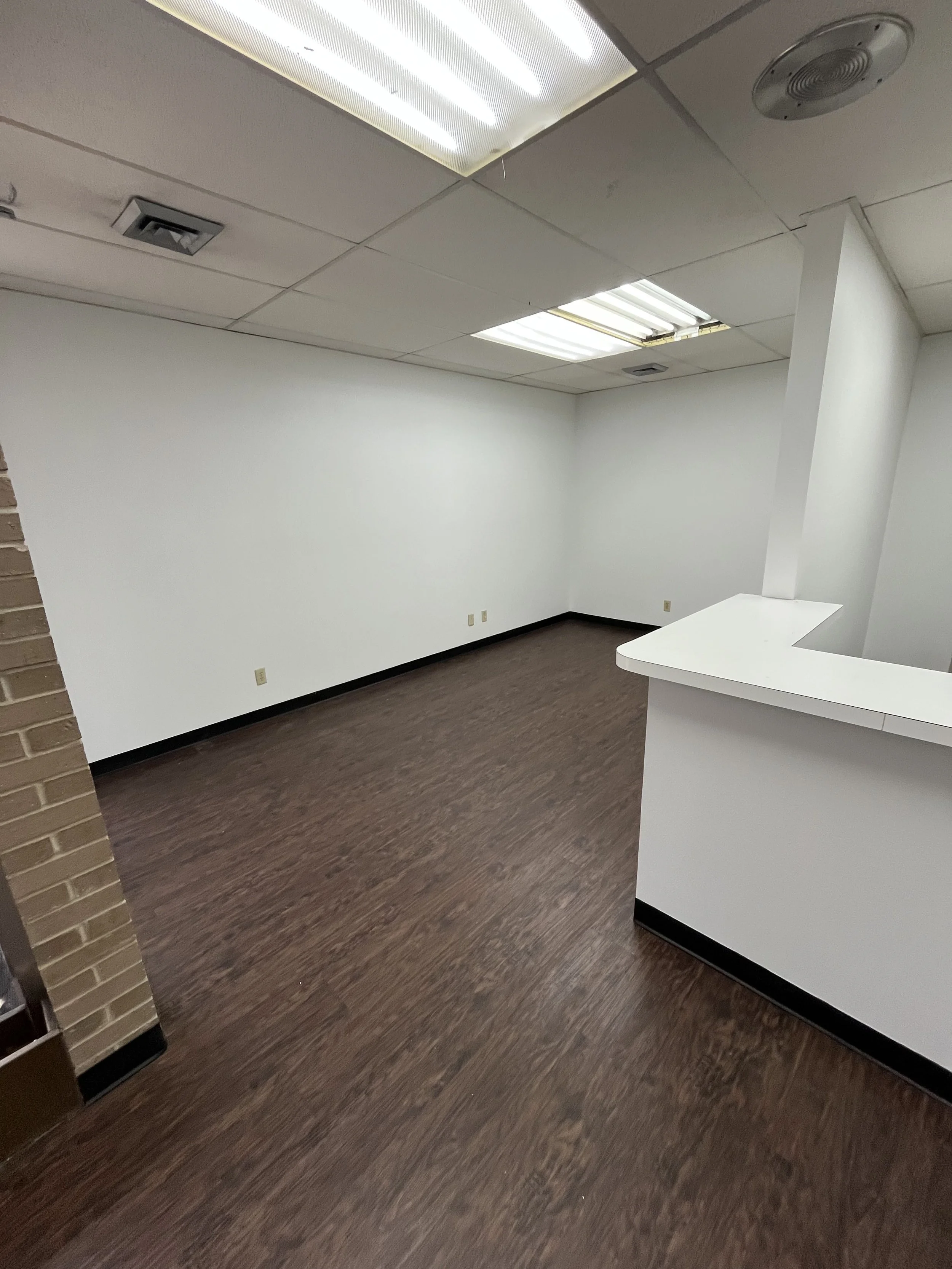 Empty office space with white walls, brown wooden floor, tile ceiling with fluorescent lighting, and a partial wall with a white countertop.