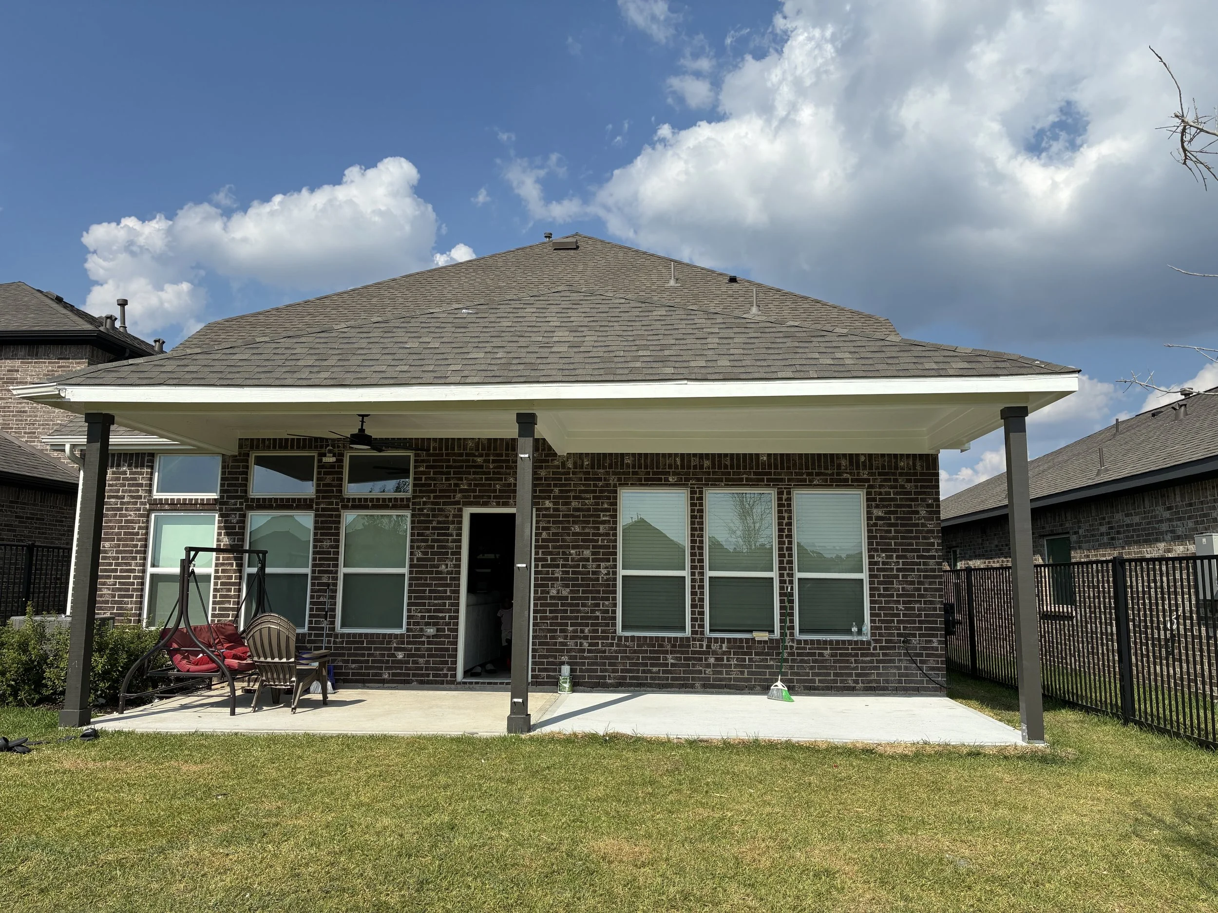 Rear view of a brick house with a covered patio, four large windows, and a door, with outdoor furniture and a green lawn under a partly cloudy sky.