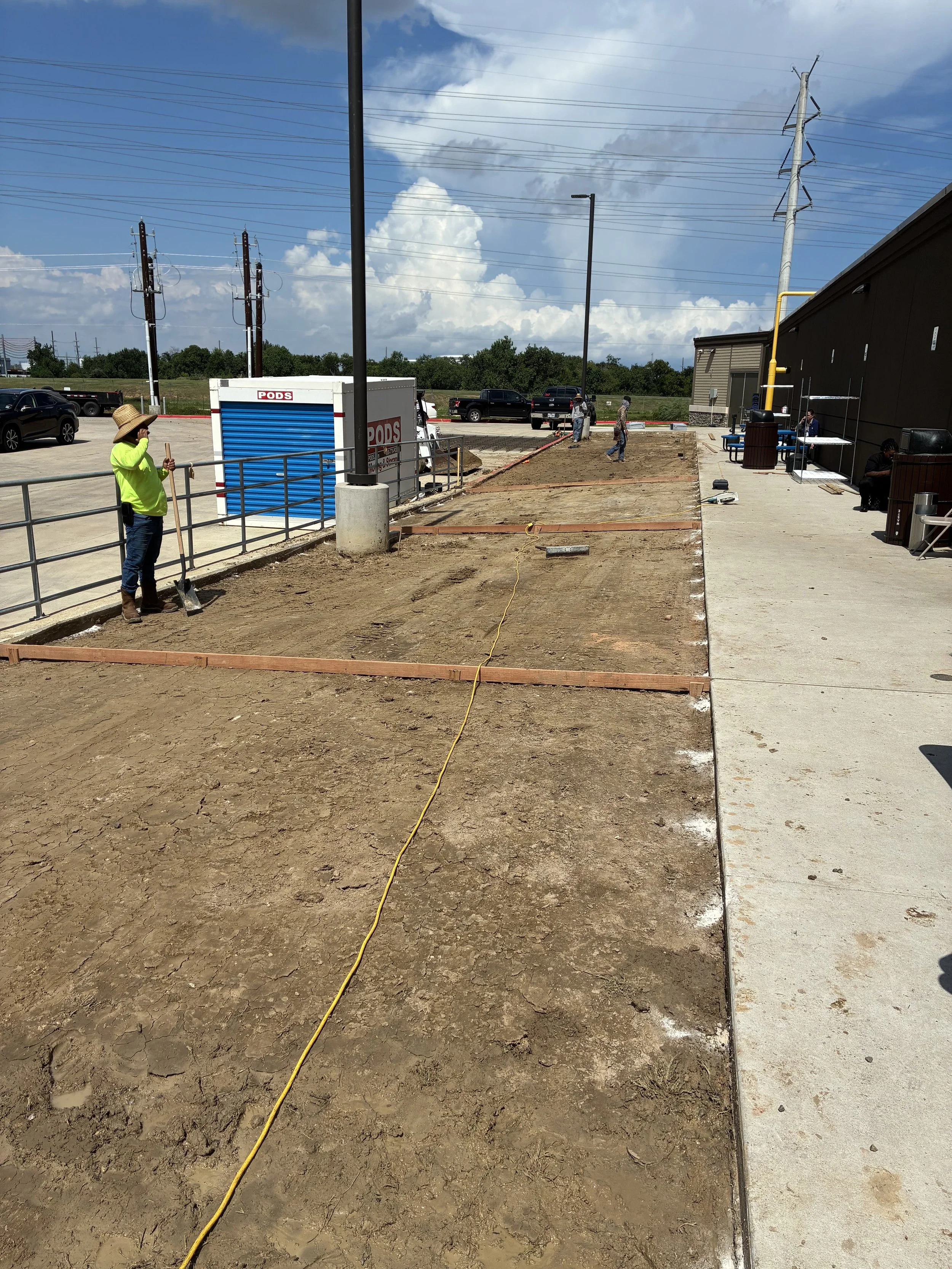 Construction workers working on a sidewalk and landscape area outside a building on a sunny day with partly cloudy sky.