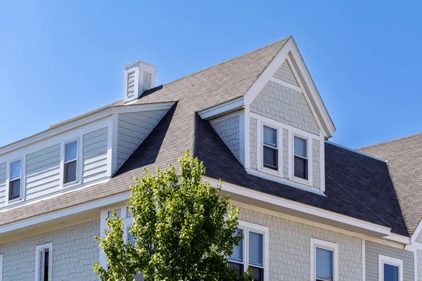 Upper portion of a white house with a steep gable roof, dormer windows, and a chimney, under a clear blue sky. A leafy green tree partially obscures the house.