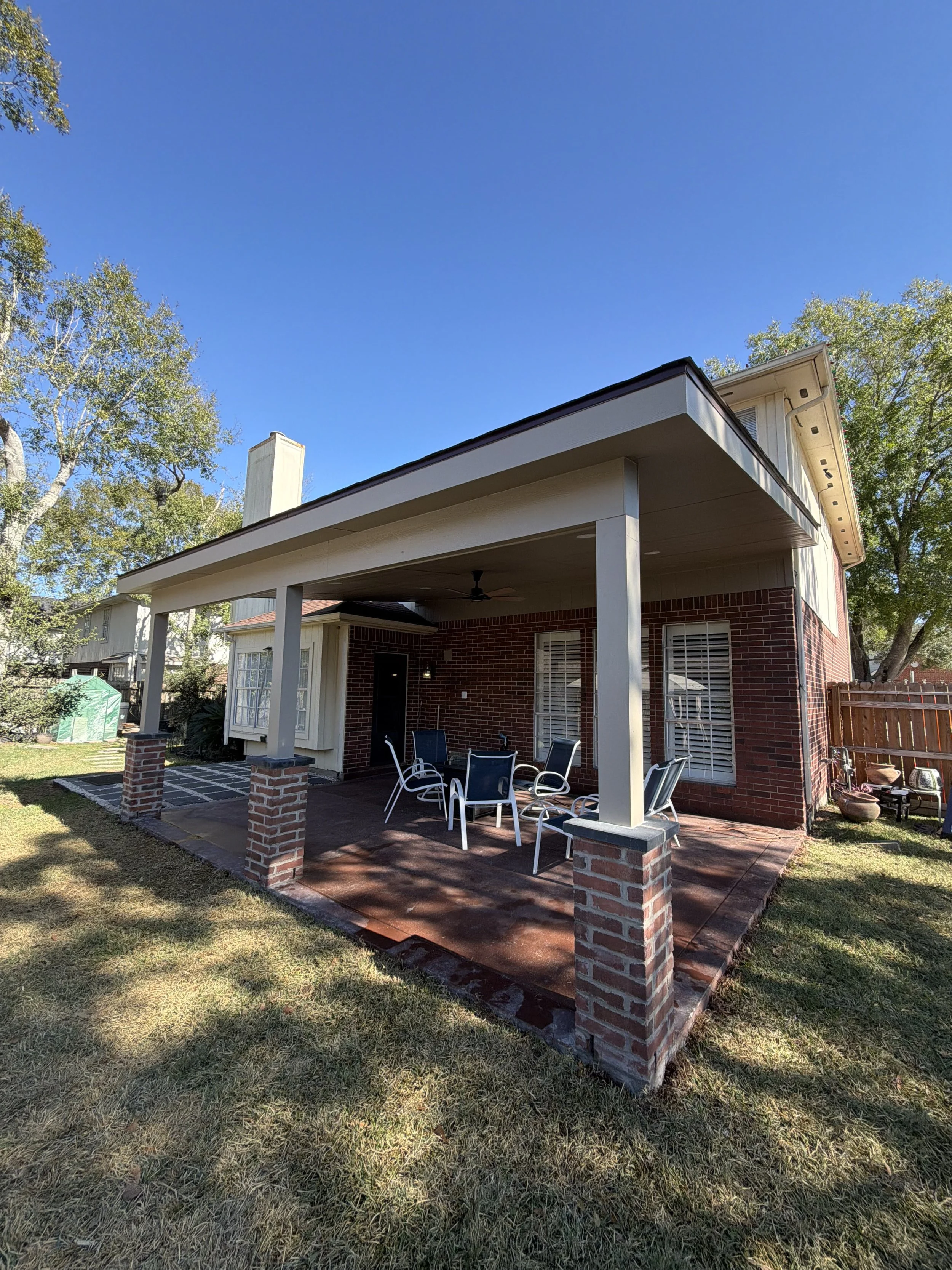 Backyard patio with a covered area, outdoor dining table, and chairs, brick and wood house exterior, green lawn, trees, and clear blue sky.