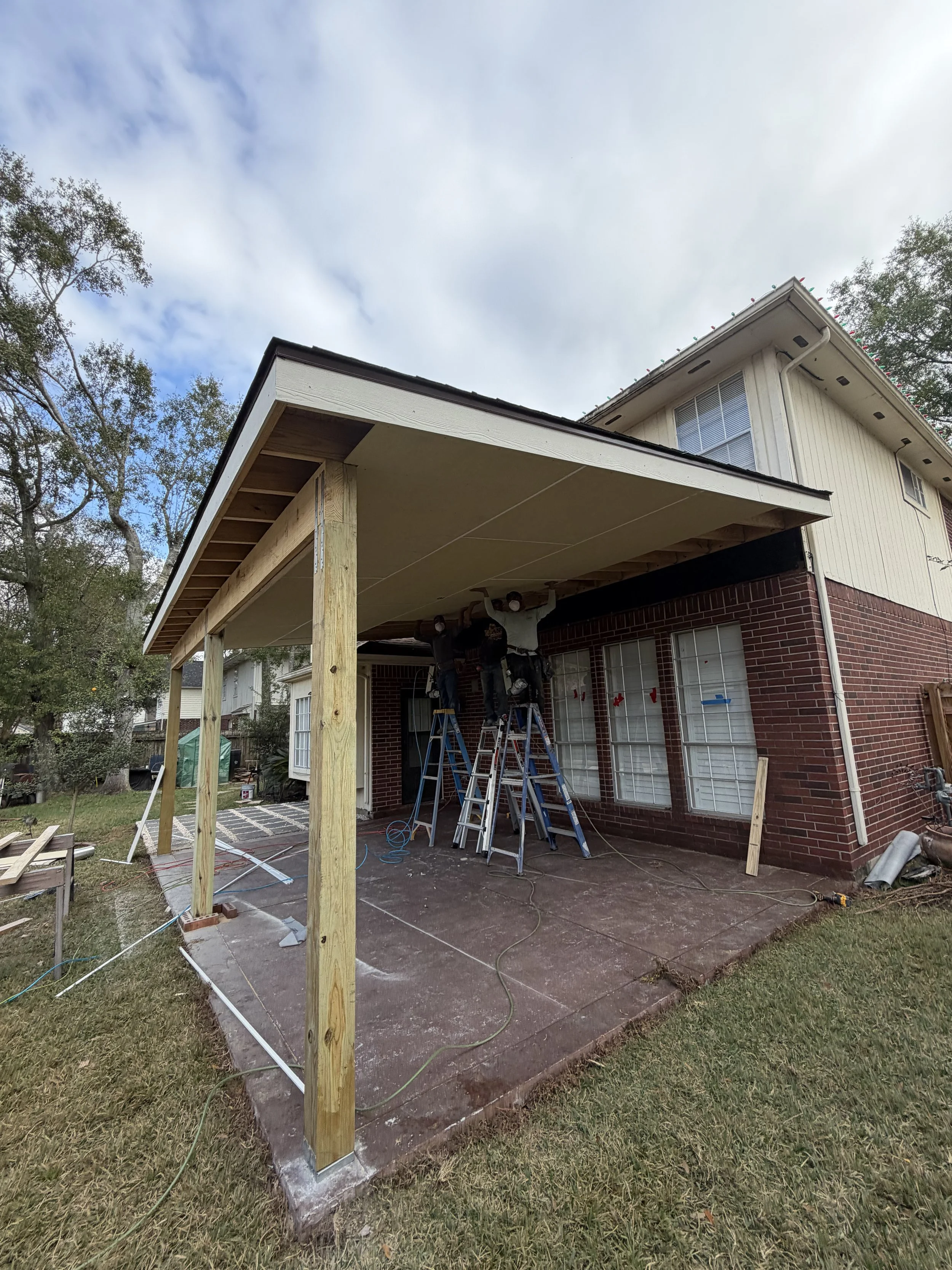 Two workers on ladders constructing a porch extension on a red brick house, with a wooden frame and white ceiling panels.