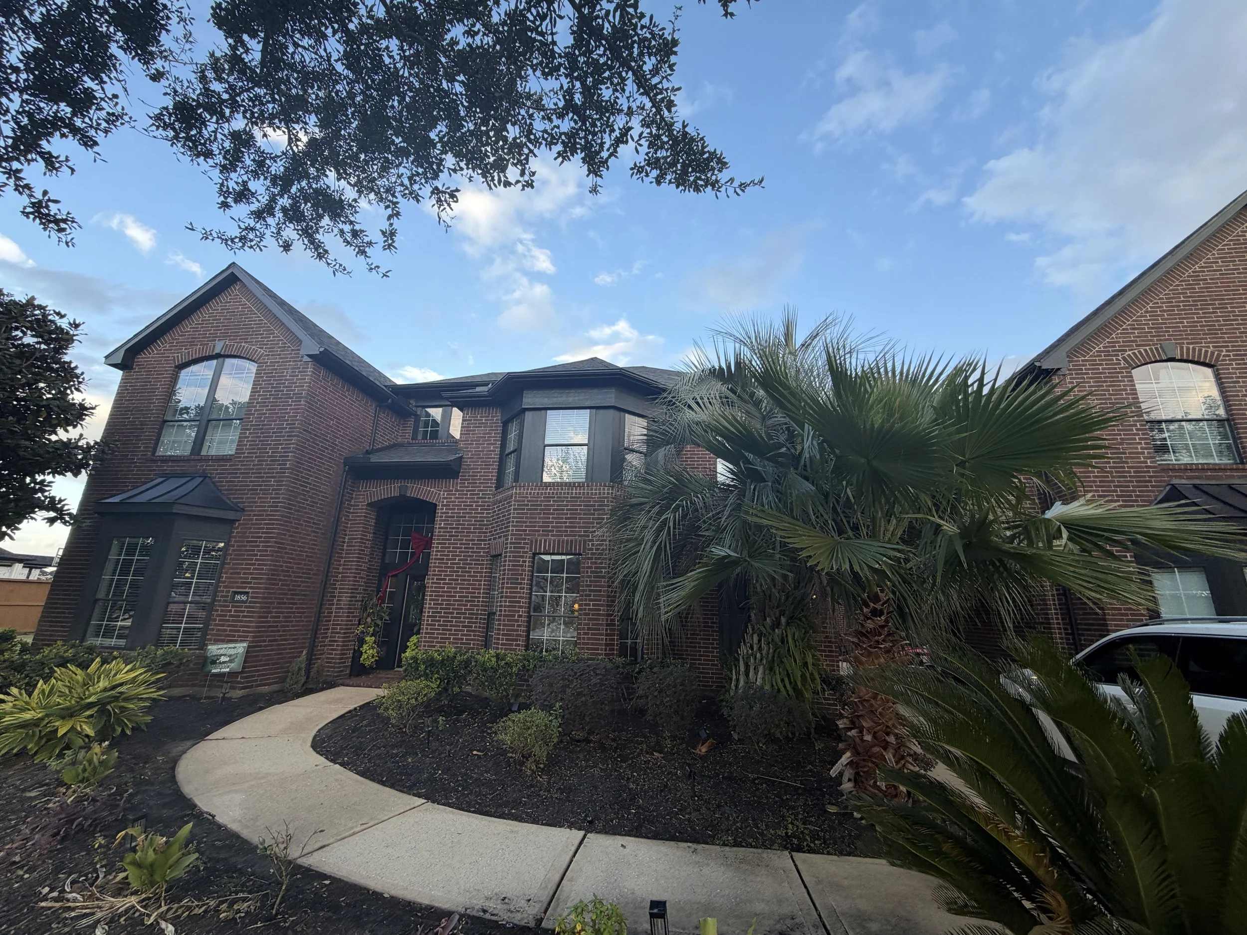 A two-story brick house with large windows and a curved pathway in the front yard, featuring a palm tree and other shrubs, under a partly cloudy sky.