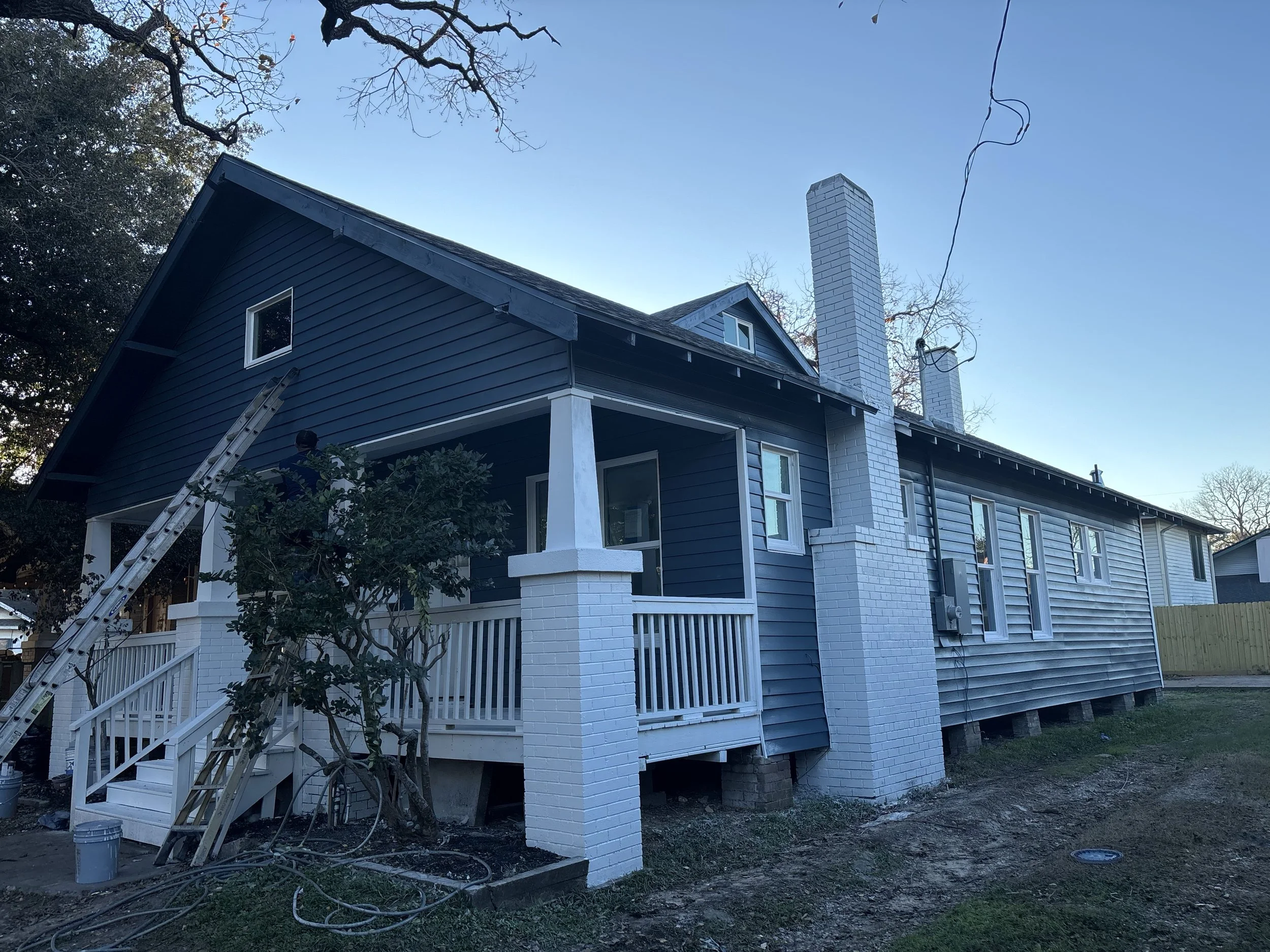 Side view of a house with dark blue siding, white chimney, and a small porch with white railing, under construction or renovation.