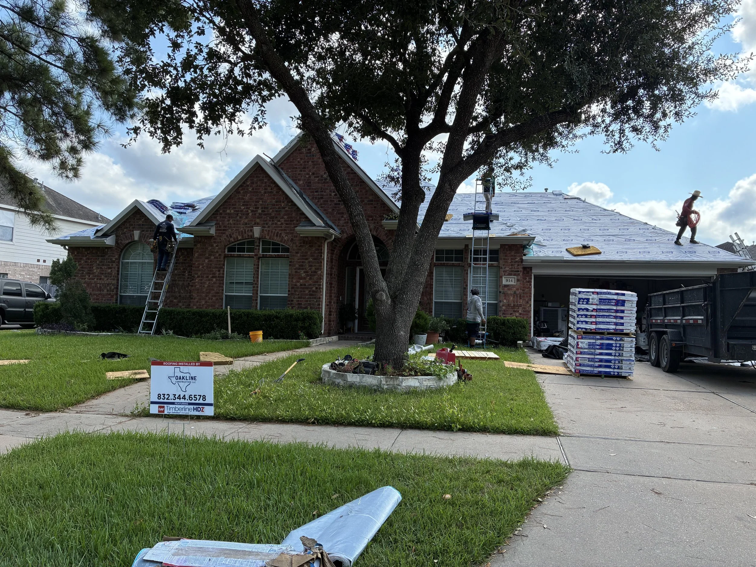 A house under construction with workers on the roof installing a new shingle roof. Some workers are using ladders and a lift to reach the roof. There is a sign on the lawn with contact information for roofing services and some construction materials 