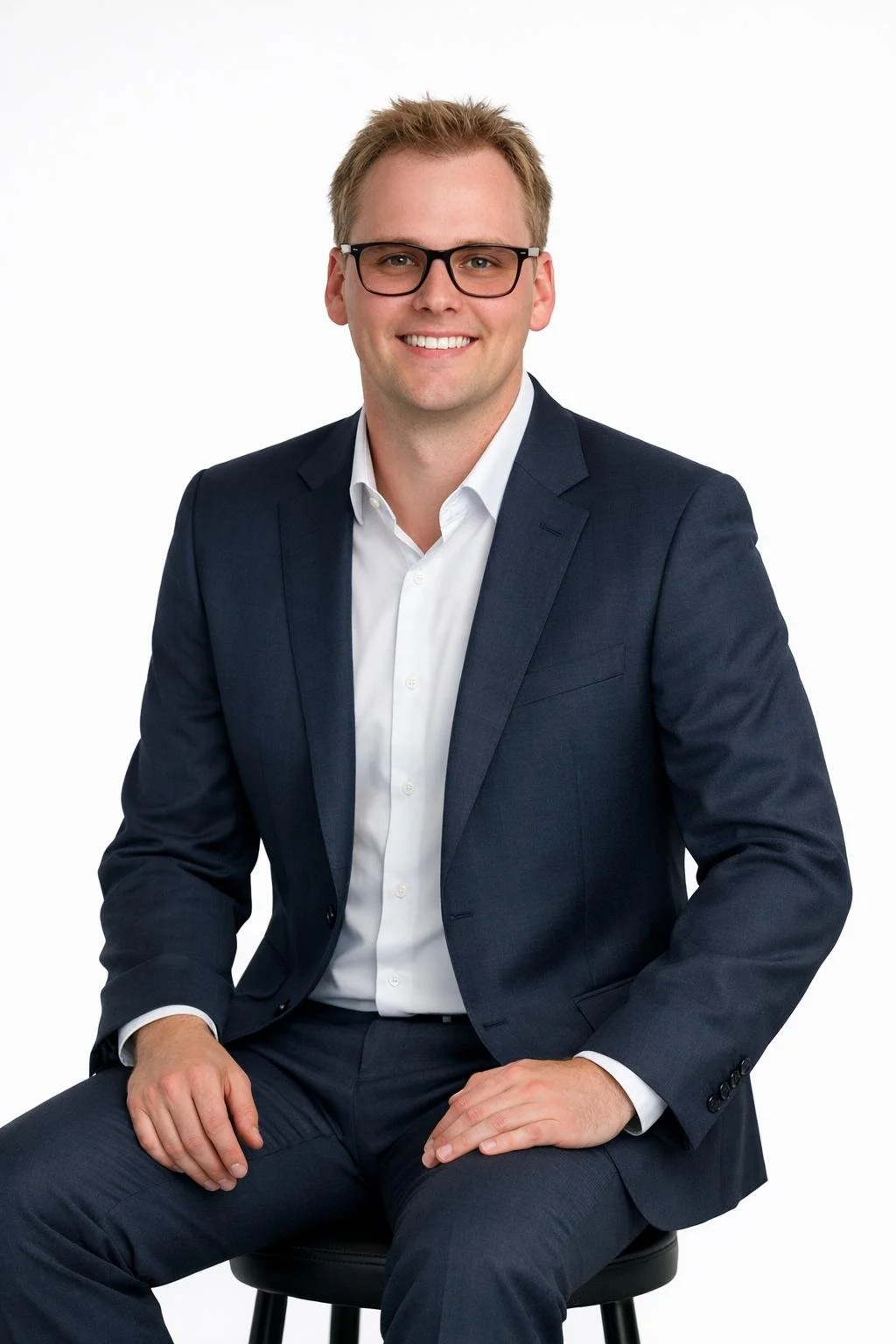 A young man with light brown hair, glasses, wearing a navy blue suit and white shirt, smiling, sitting on a black chair against a plain white background.