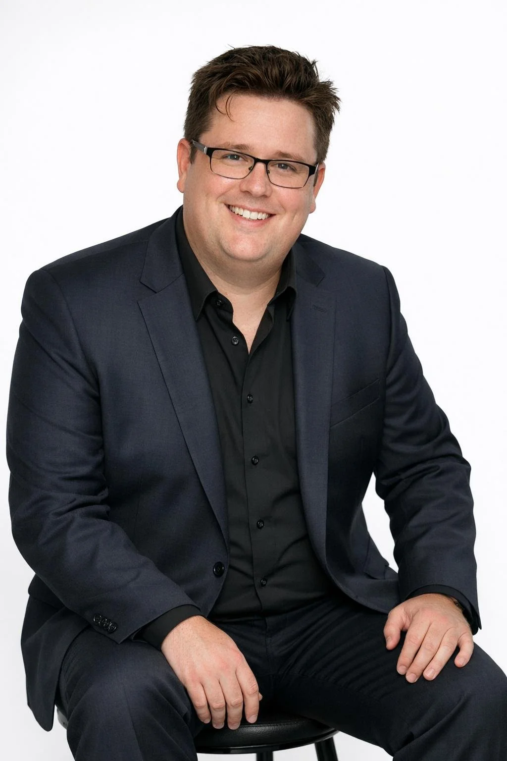 A smiling man in a black suit and glasses sitting on a stool against a white background.