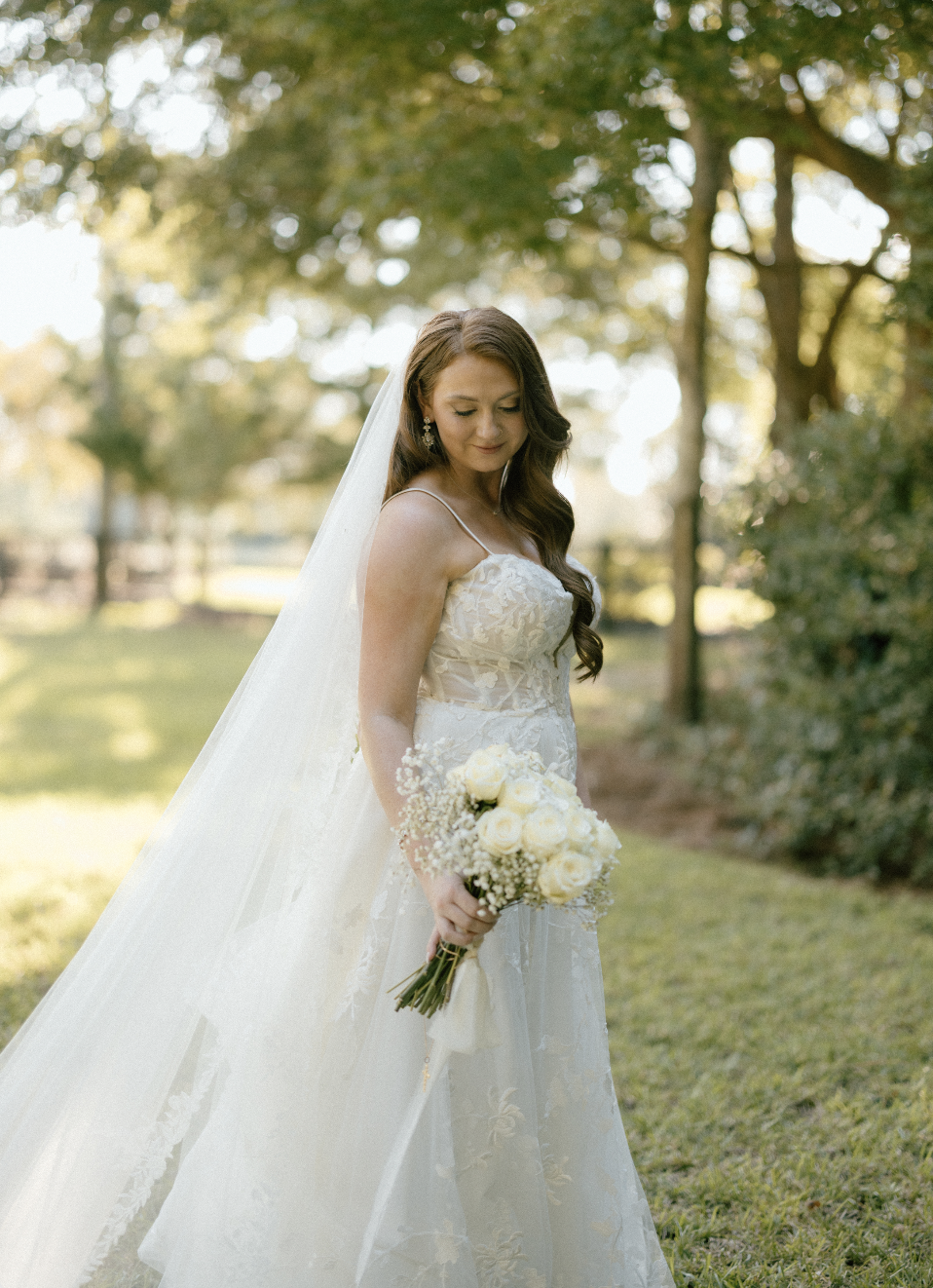 A bride standing outdoors in a garden, holding a bouquet of white roses and baby's breath, wearing a lace wedding gown and veil, with trees and sunlight in the background.