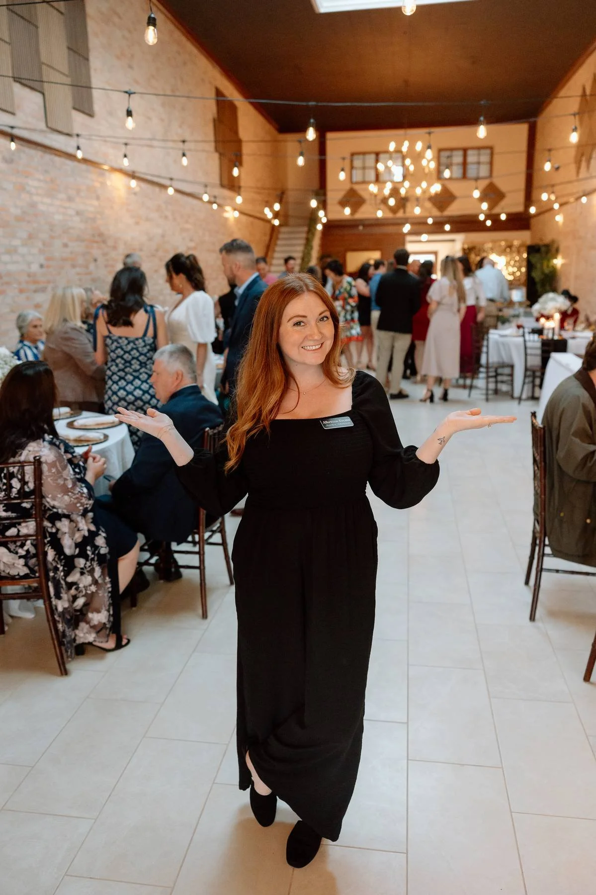 A woman in a black dress with a name tag standing with her arms outstretched in a decorated banquet hall, smiling at the camera. Guests are mingling and dining in the background at a venue with string lights and a chandelier.