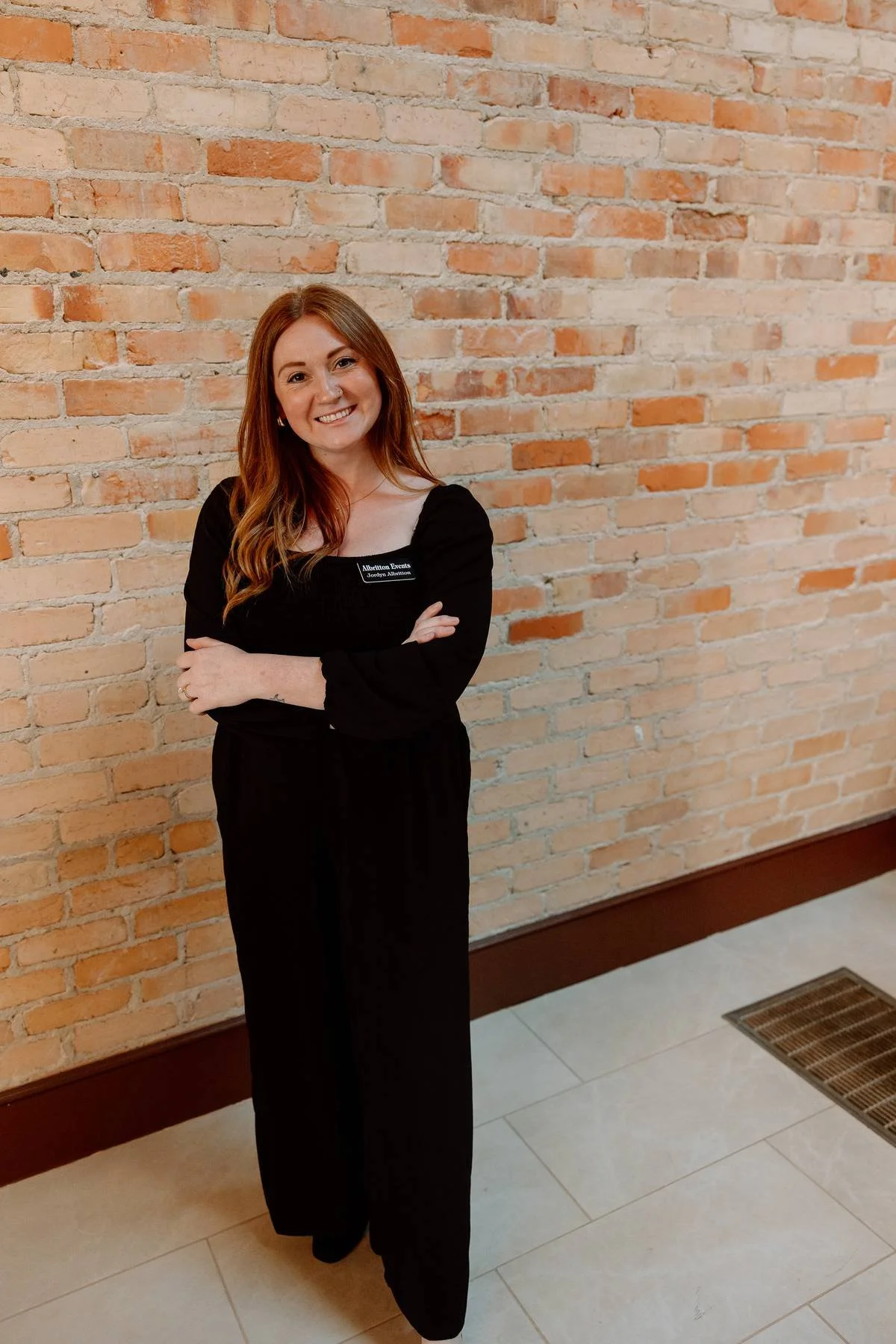 A woman with red hair, wearing black attire with a name tag, standing against a brick wall in an indoor setting.