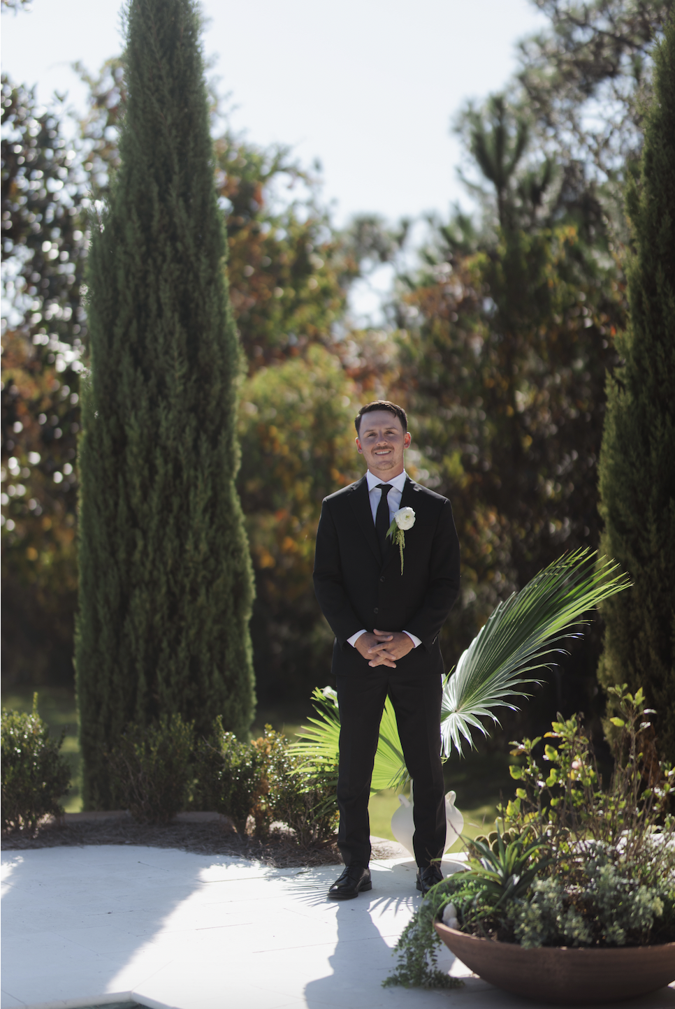 A man in a black suit with a white shirt and black tie, wearing a white boutonnière, standing outdoors on a bright day surrounded by tall cypress trees and tropical plants.