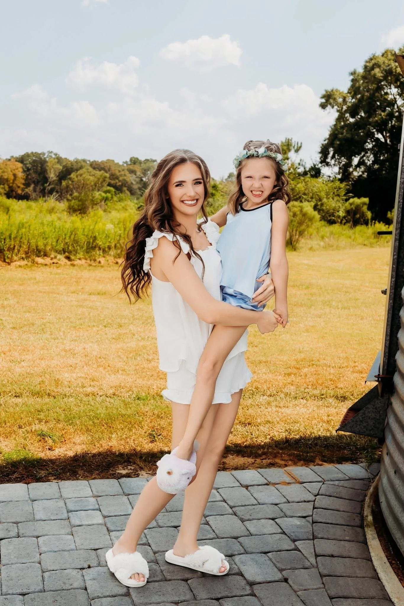 A young woman holding a little girl in her arms outdoors on a sunny day, with a green field and trees in the background, smiling at the camera.