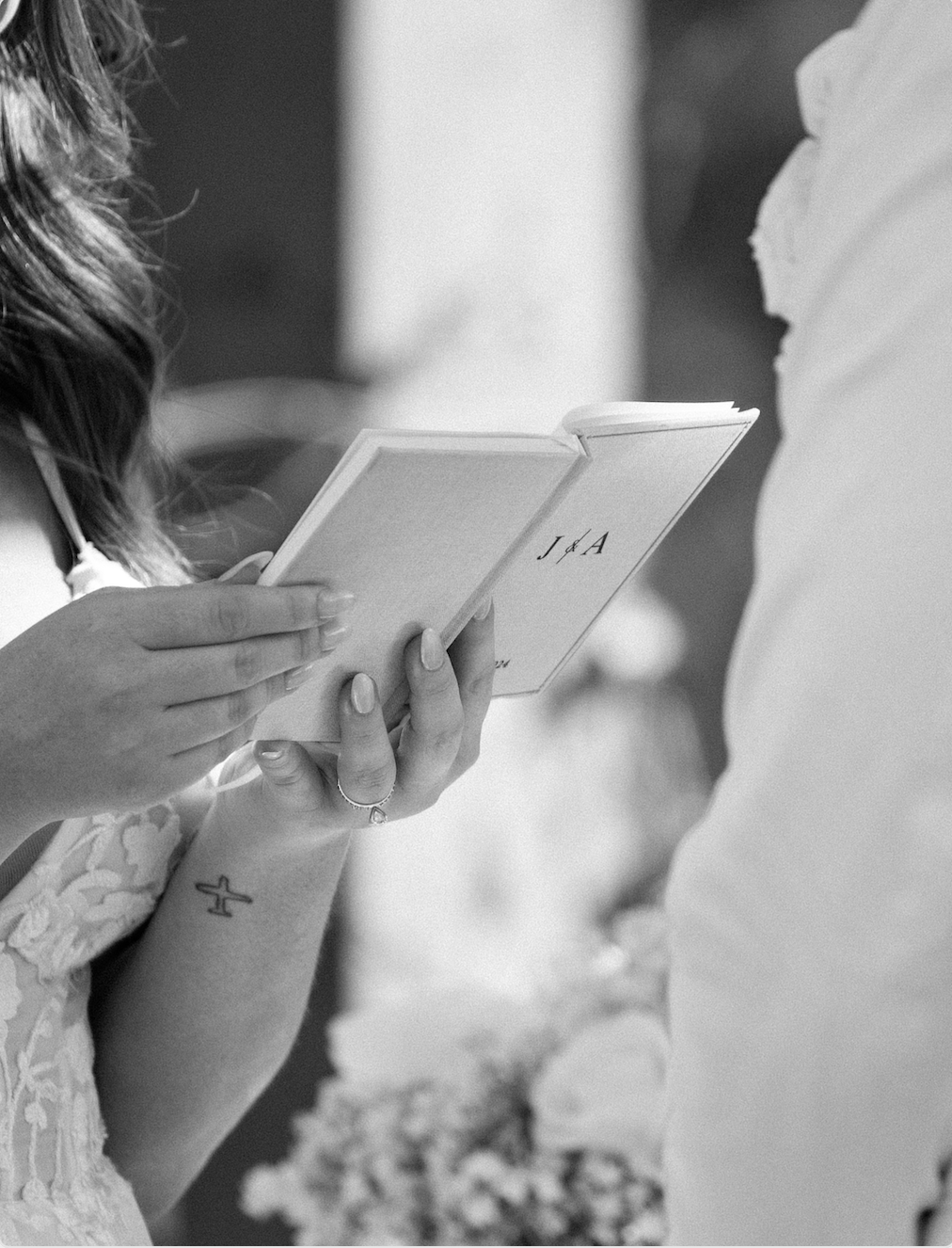 A person holding a small book or card, possibly during a wedding or special event, with a woman wearing a lace dress and a tattoo of a cross on her wrist.