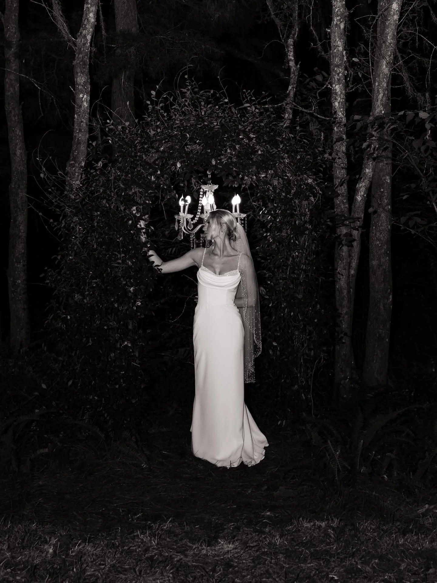 A woman in a white wedding dress standing outdoors at night with a chandelier above her head, surrounded by trees.
