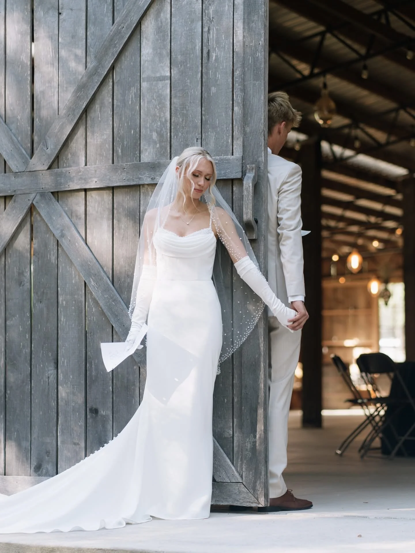 A bride and groom holding hands behind a rustic wooden barn door, with the bride in a white wedding gown and veil, and the groom in a white tuxedo, during a wedding ceremony.