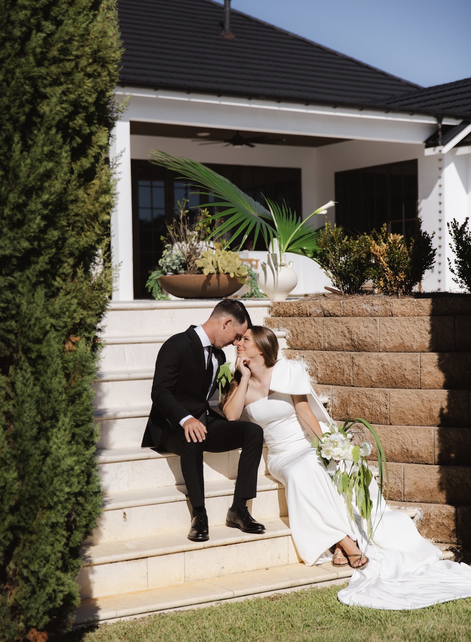 Bride and groom sitting on outdoor steps, touching foreheads, with wedding dress and suit, in front of house with potted plants.