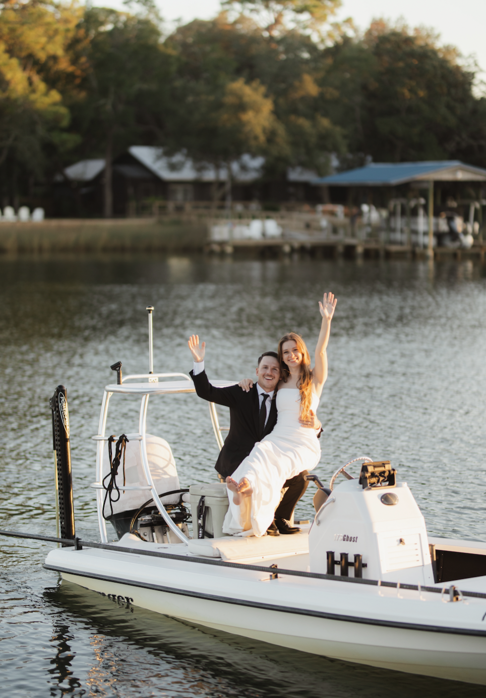 A newlywed couple in wedding attire celebrating on a boat with a lake and wooded shoreline in the background.