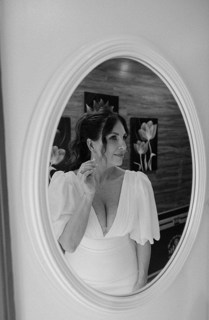 Black and white photo of a woman looking at her reflection in an oval mirror, touching her earring, with a wooden wall and floral art in the background.