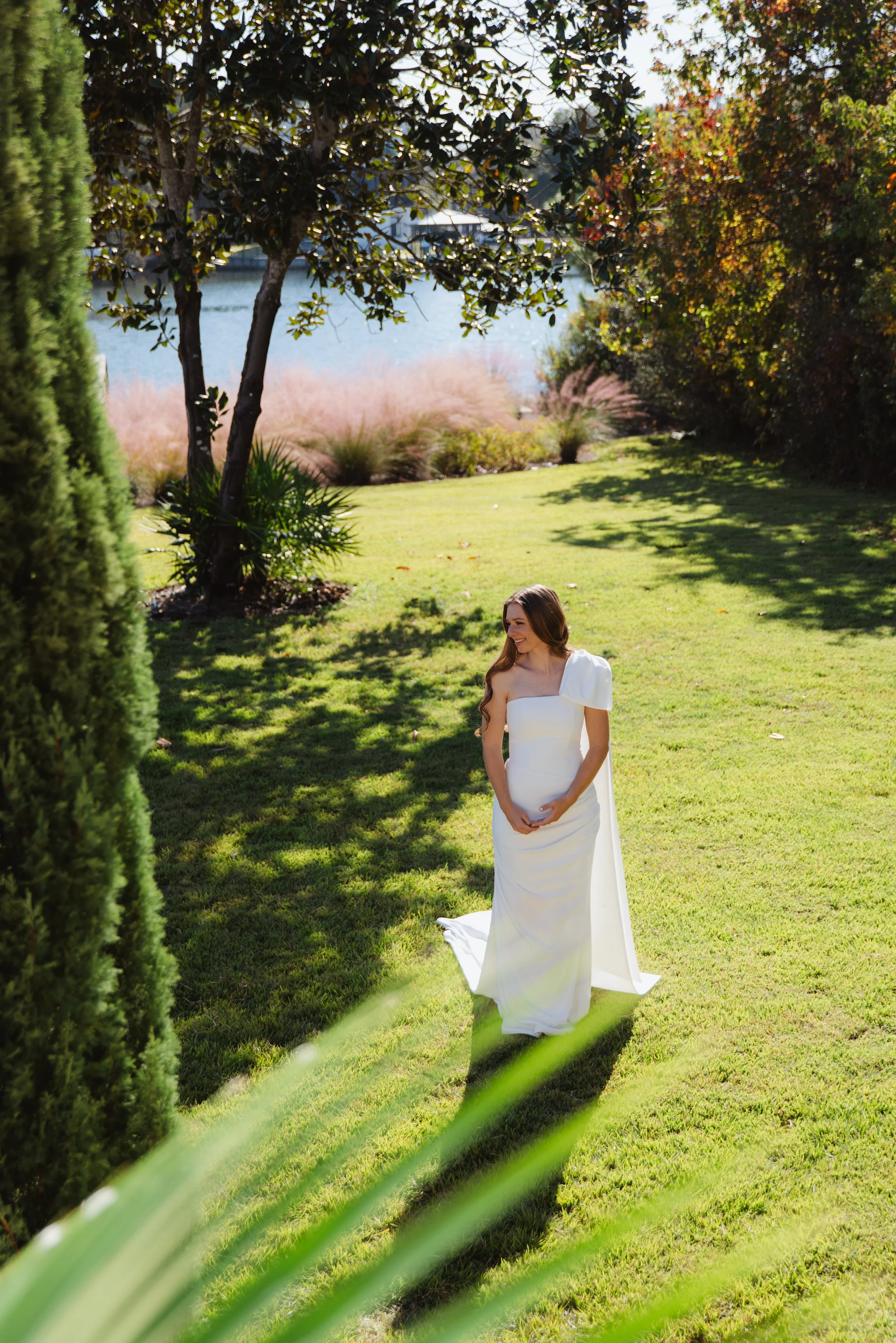 A woman in a white dress stands on a green lawn near trees, with water and a dock in the background, sunlight casting shadows.