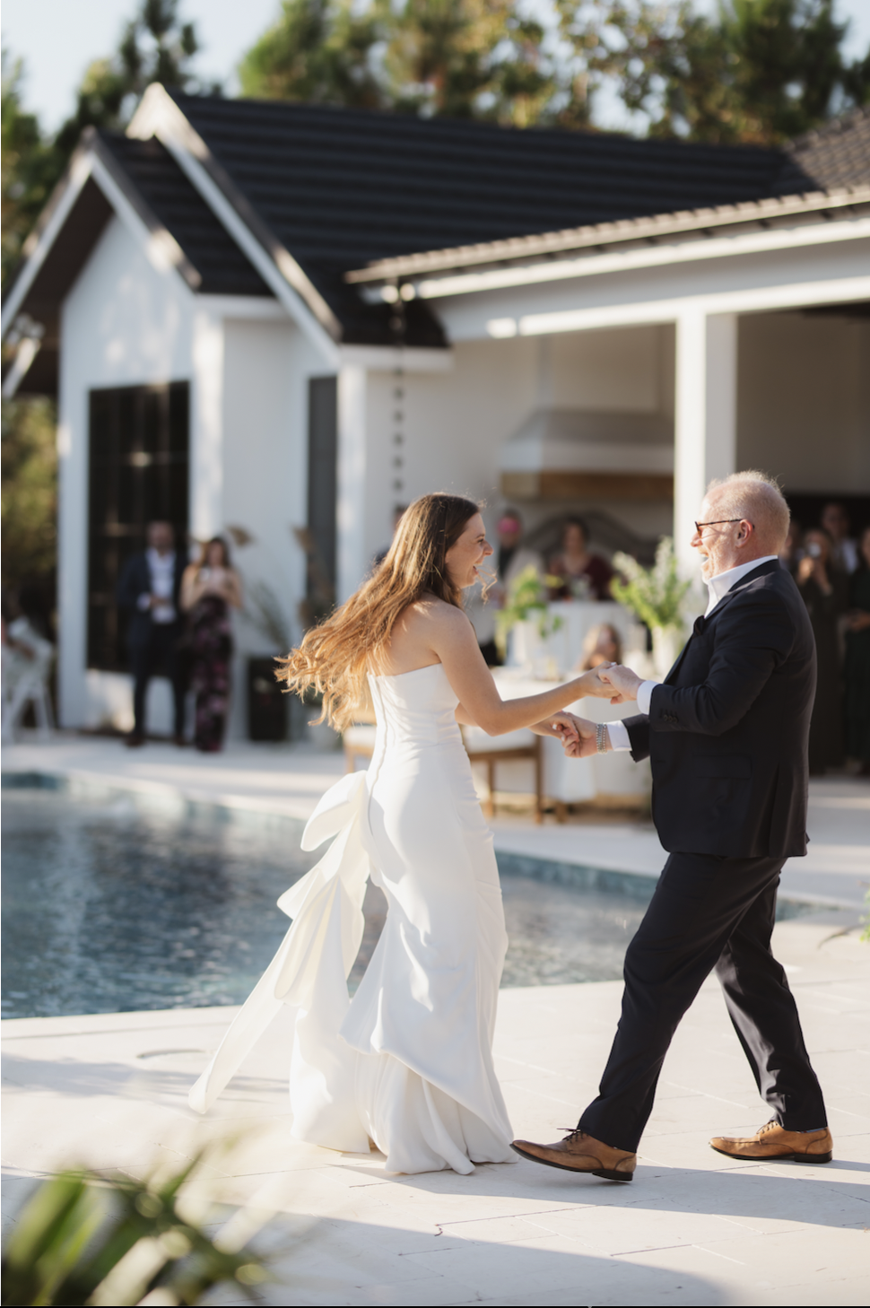 A bride and an older man dancing at an outdoor wedding reception by a pool, with guests in the background.