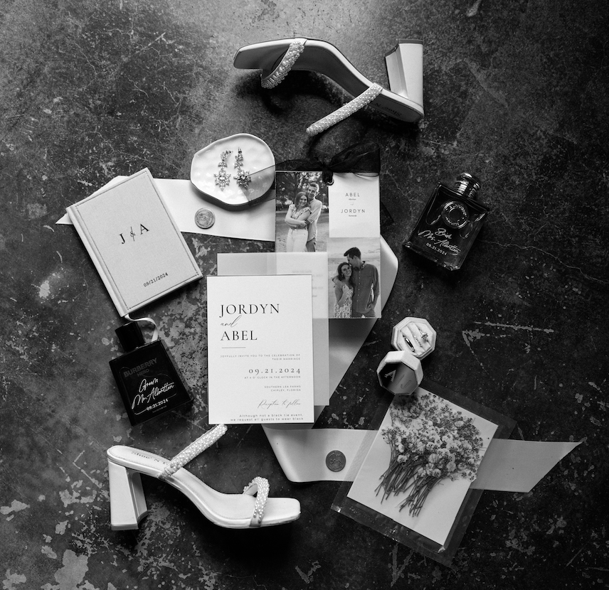 Wedding invitation, jewelry, perfume, high heel shoes, photographs, and other wedding accessories on a dark surface.