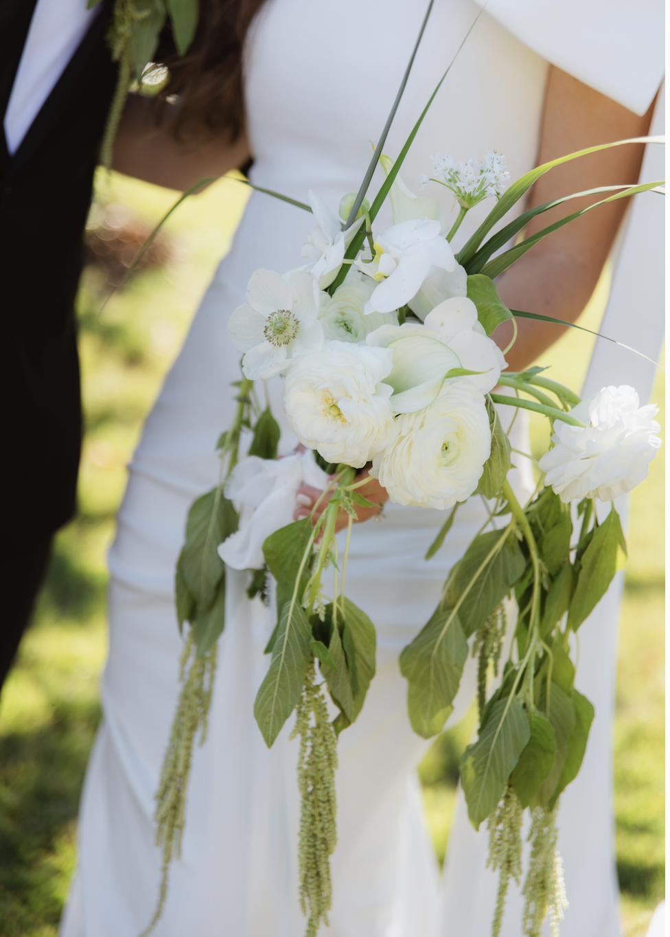 A bride in a white dress holds a cascading bouquet of white flowers and green foliage at an outdoor wedding.