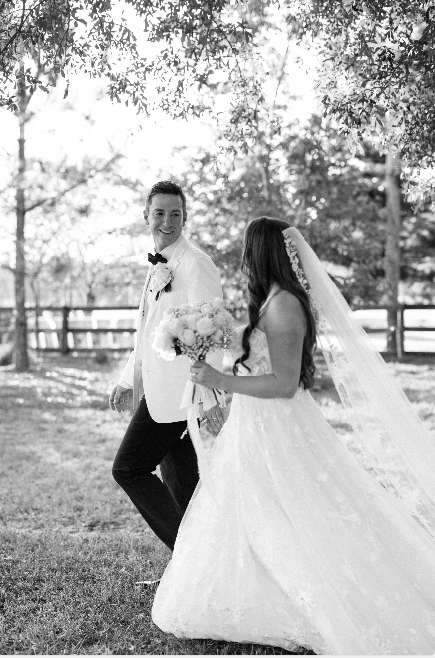 A black-and-white photo of a bride and groom outdoors on their wedding day, holding hands and smiling at each other, with trees and a wooden fence in the background.