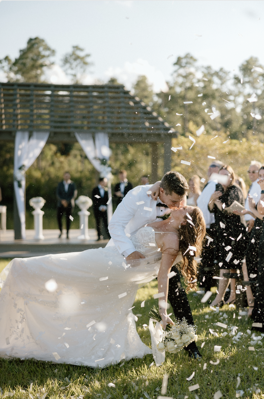 A newlywed couple is dancing outdoors at their wedding, with the groom dipping the bride, surrounded by confetti and guests, during the day.
