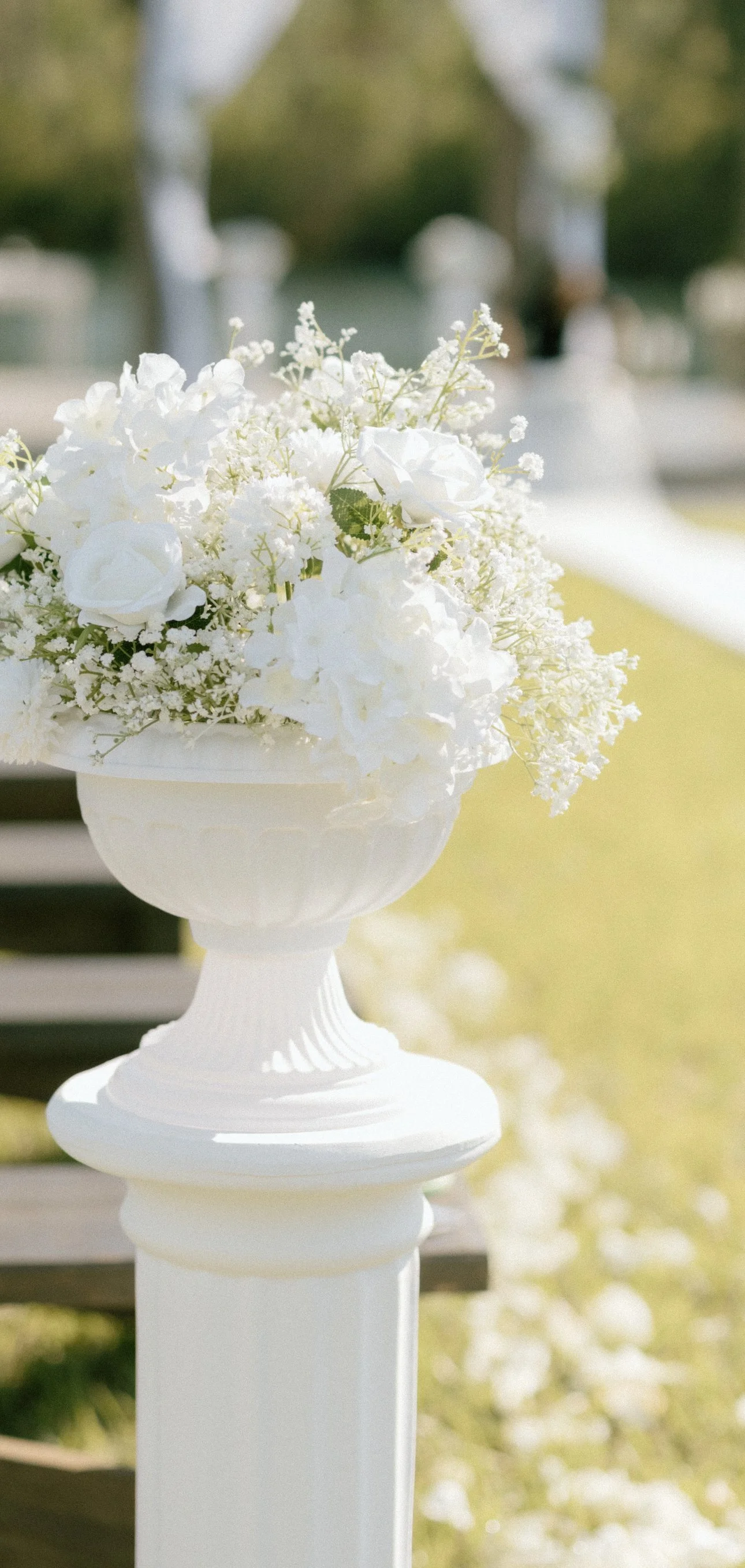 White floral arrangement in a white vase on a pedestal, set outdoors with a blurred background.
