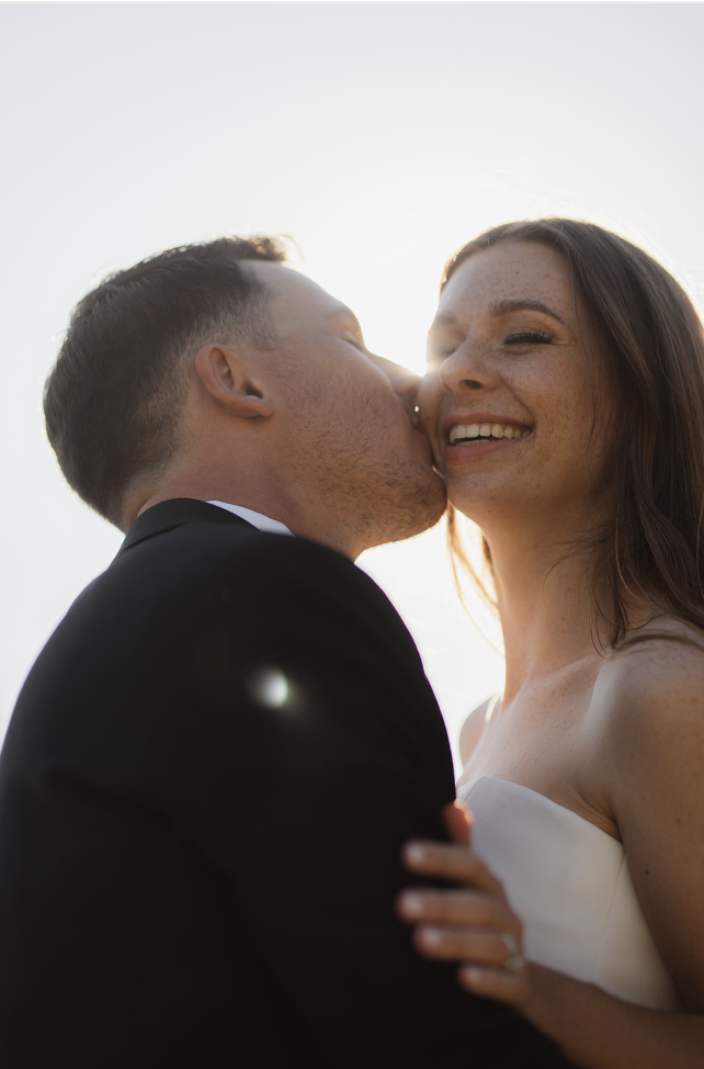 A newlywed couple shares a kiss outdoors, with the man in a black suit and the woman in a white dress, illuminated by sunlight behind them.