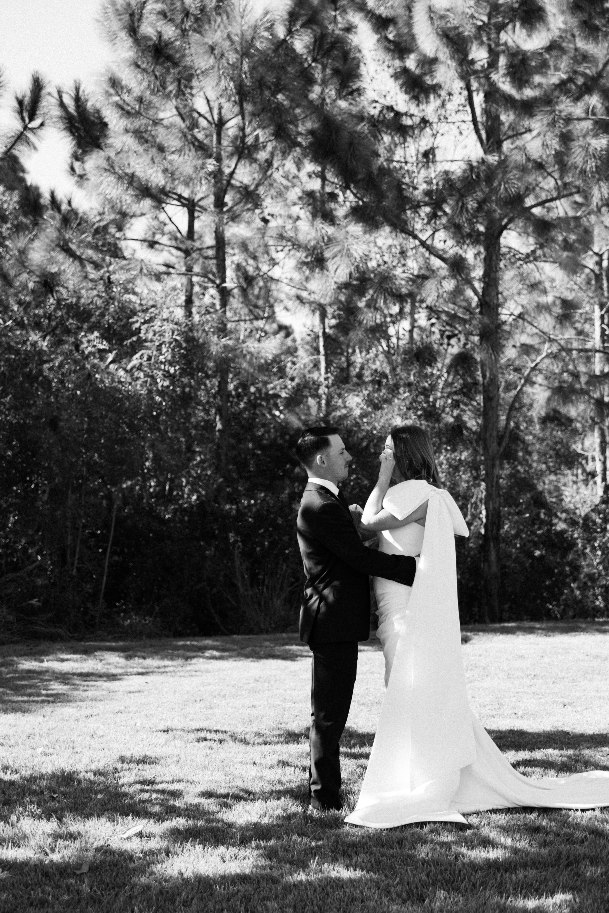 A black-and-white photo of a bride and groom standing outdoors, facing each other, surrounded by tall trees and grass.