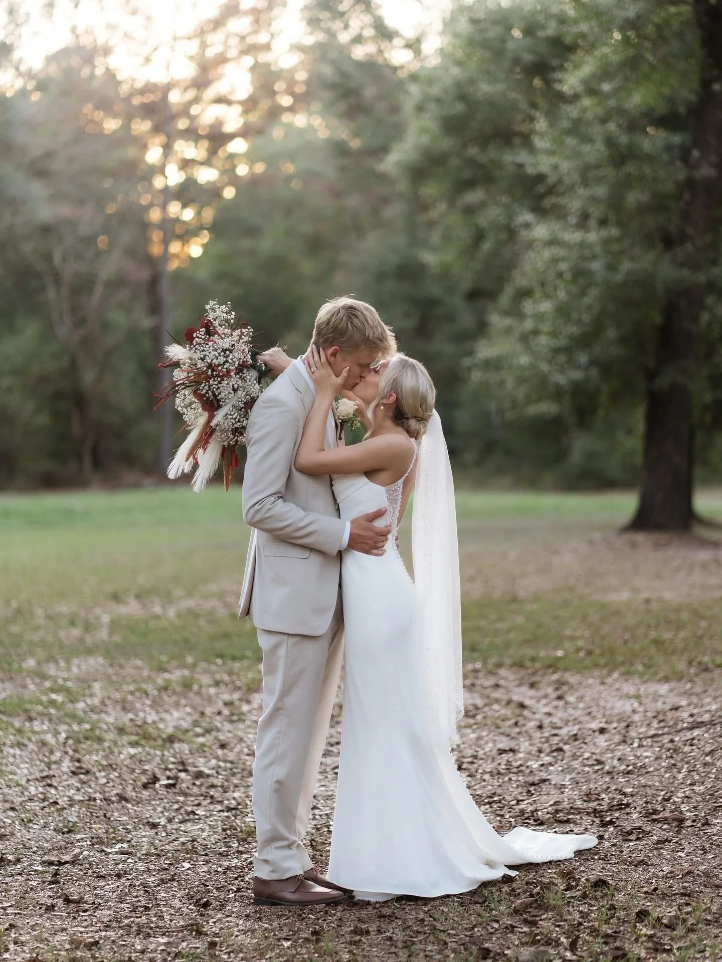 A couple in wedding attire sharing a kiss outside in a wooded area with sunlight filtering through trees.