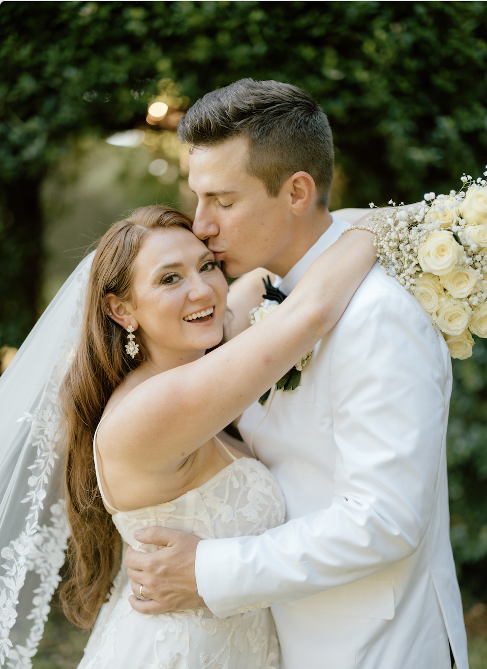 A newlywed couple, the man in a white tuxedo with a black bowtie, kisses the bride on her forehead. The bride has long red hair, wears a lace wedding dress, earrings, and a veil, and she is smiling with her arms around the groom. The groom has short dark hair and is holding a bouquet of white roses and baby's breath. They are outside with green trees in the background.