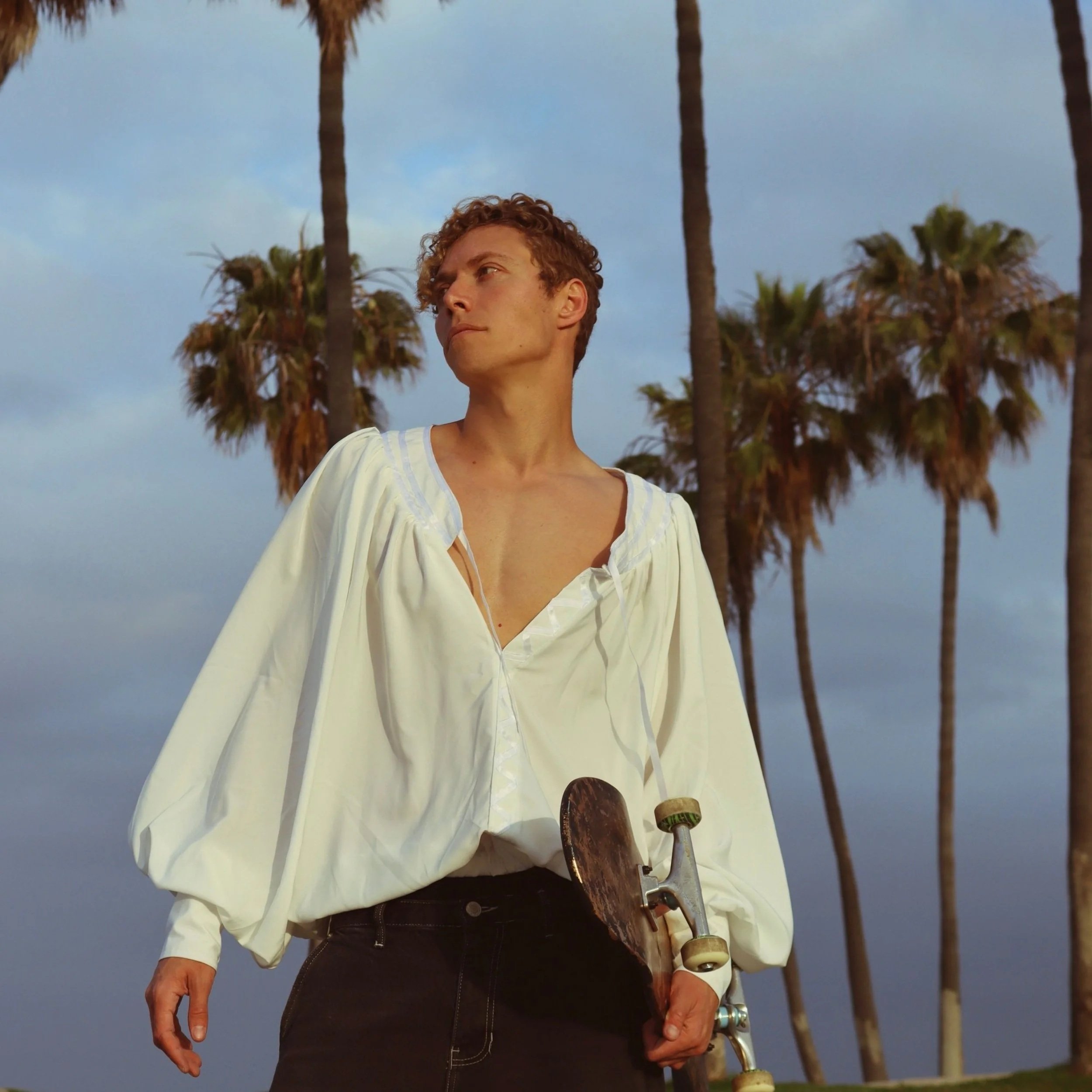 A person with curly hair stands outdoors among tall palm trees, holding a skateboard. They wear a loose, white, partially unbuttoned blouse and dark pants, looking off into the distance with a serious expression.