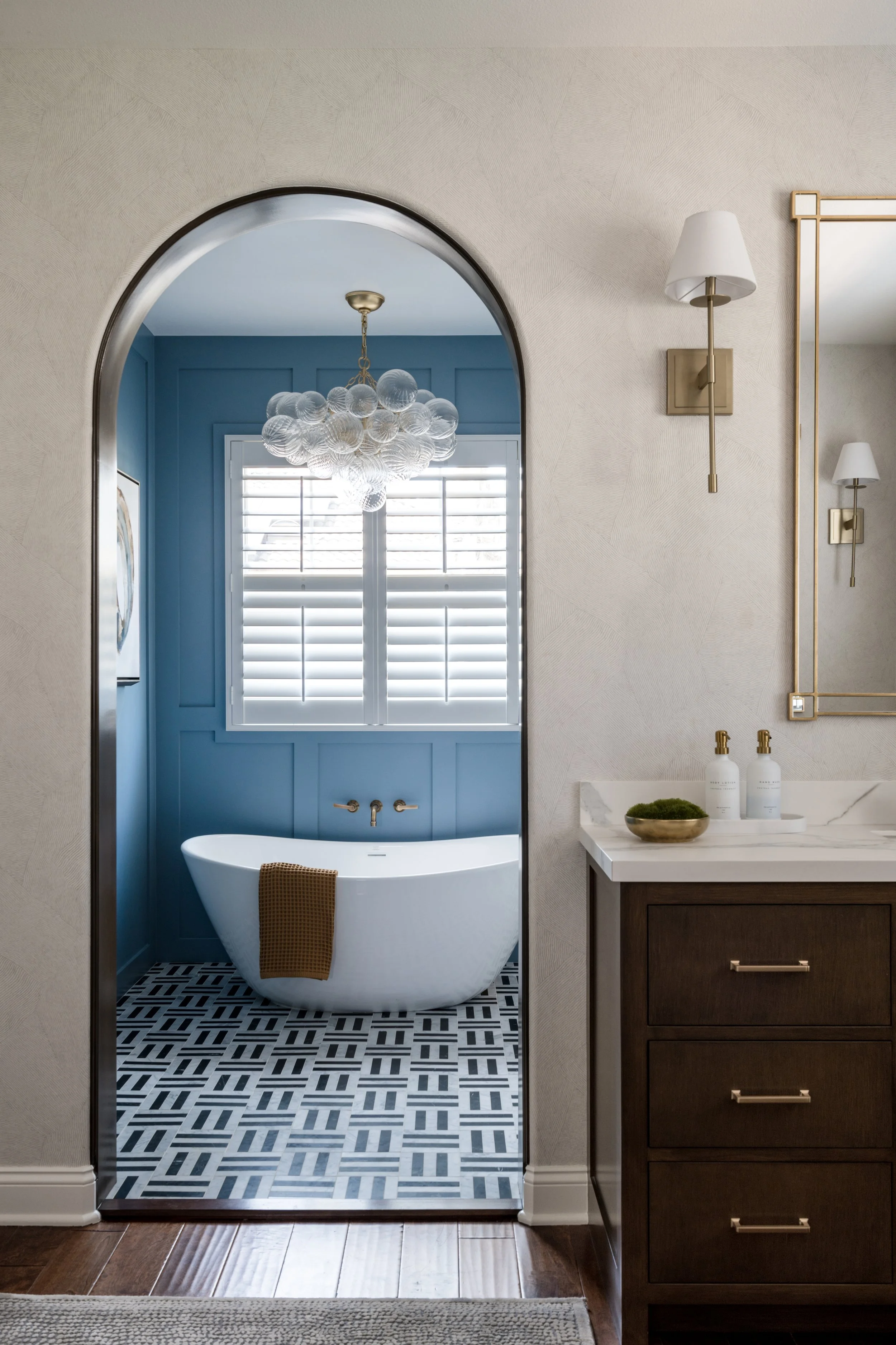 Modern bathroom with a white freestanding bathtub, blue wall paneling, black and white geometric patterned floor, and large window with white blinds, seen through an arched doorway.