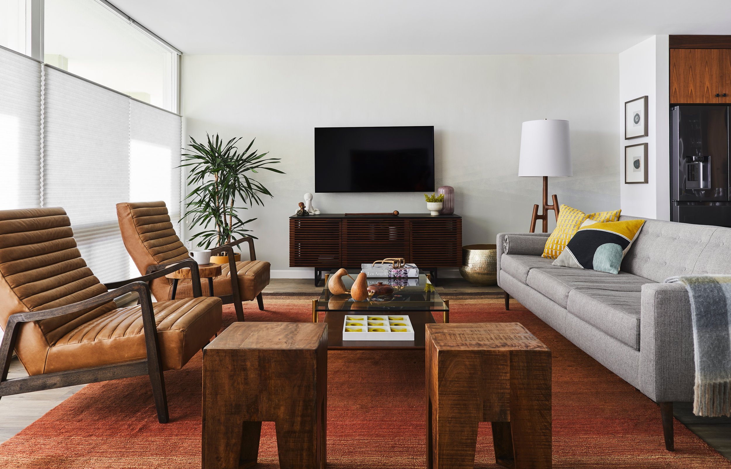 Modern living room with brown and tan chairs, gray sofa, wooden side tables, a glass coffee table, a red rug, a large plant, a wall-mounted TV, a white lamp, and decorative pillows.