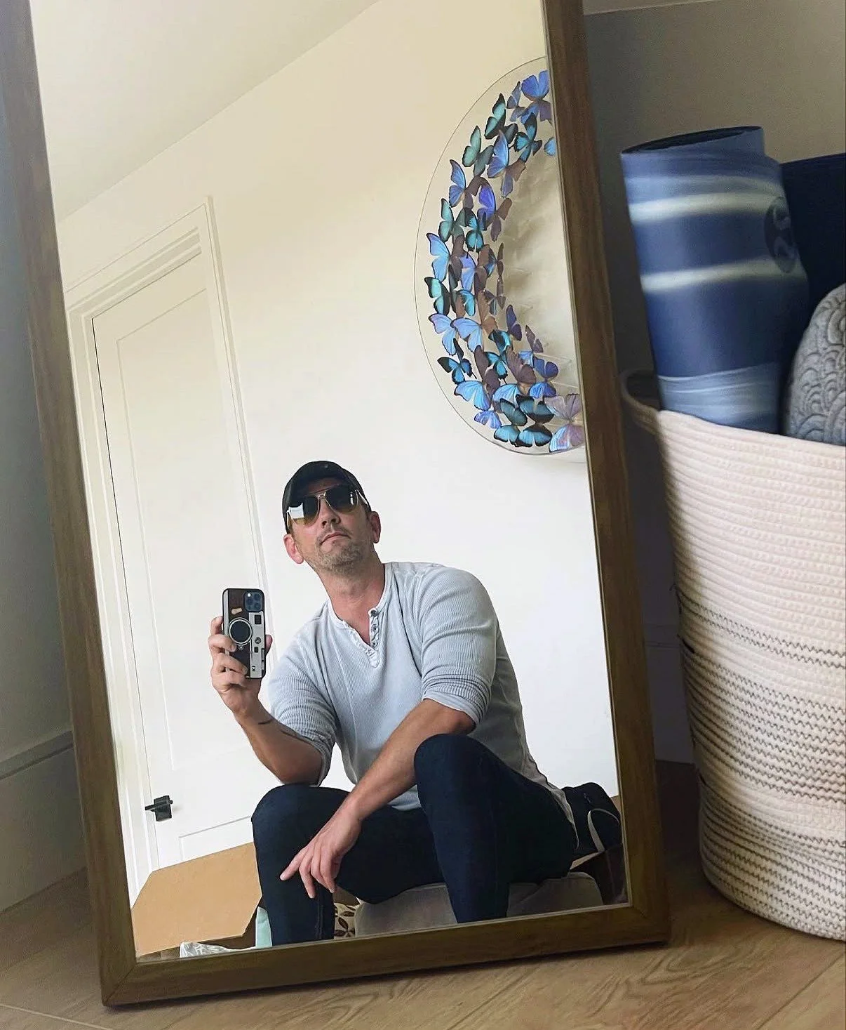 A man taking a selfie in a mirror, wearing sunglasses, a cap, a gray long-sleeve shirt, and dark pants, sitting with one knee bent. The room has a white door and a colorful wall decoration of butterflies in the background.