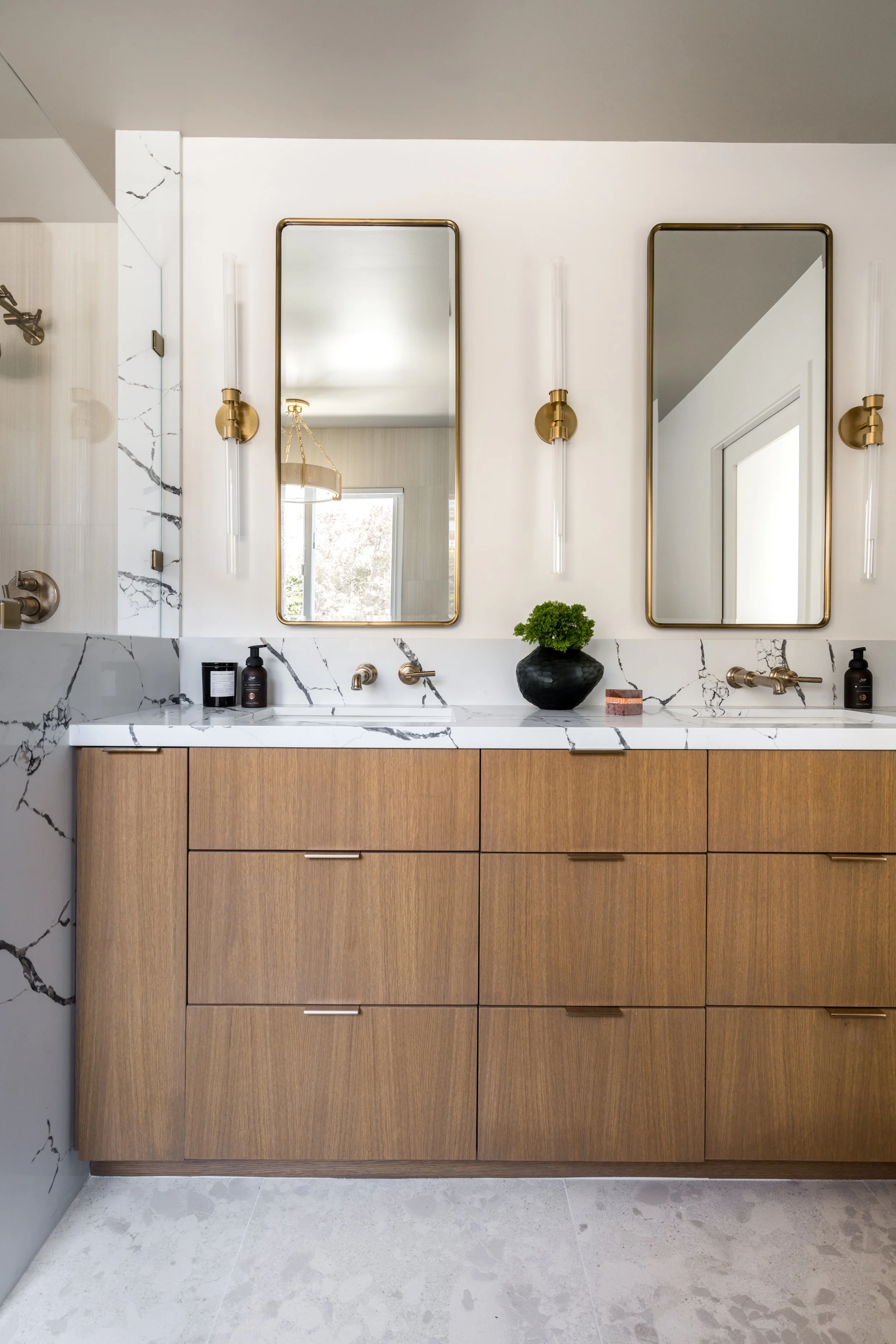 Modern bathroom vanity with two rectangular mirrors, white marble countertop with black veining, wooden cabinetry, potted plant, and black soap dispensers, illuminated by vertical sconces.