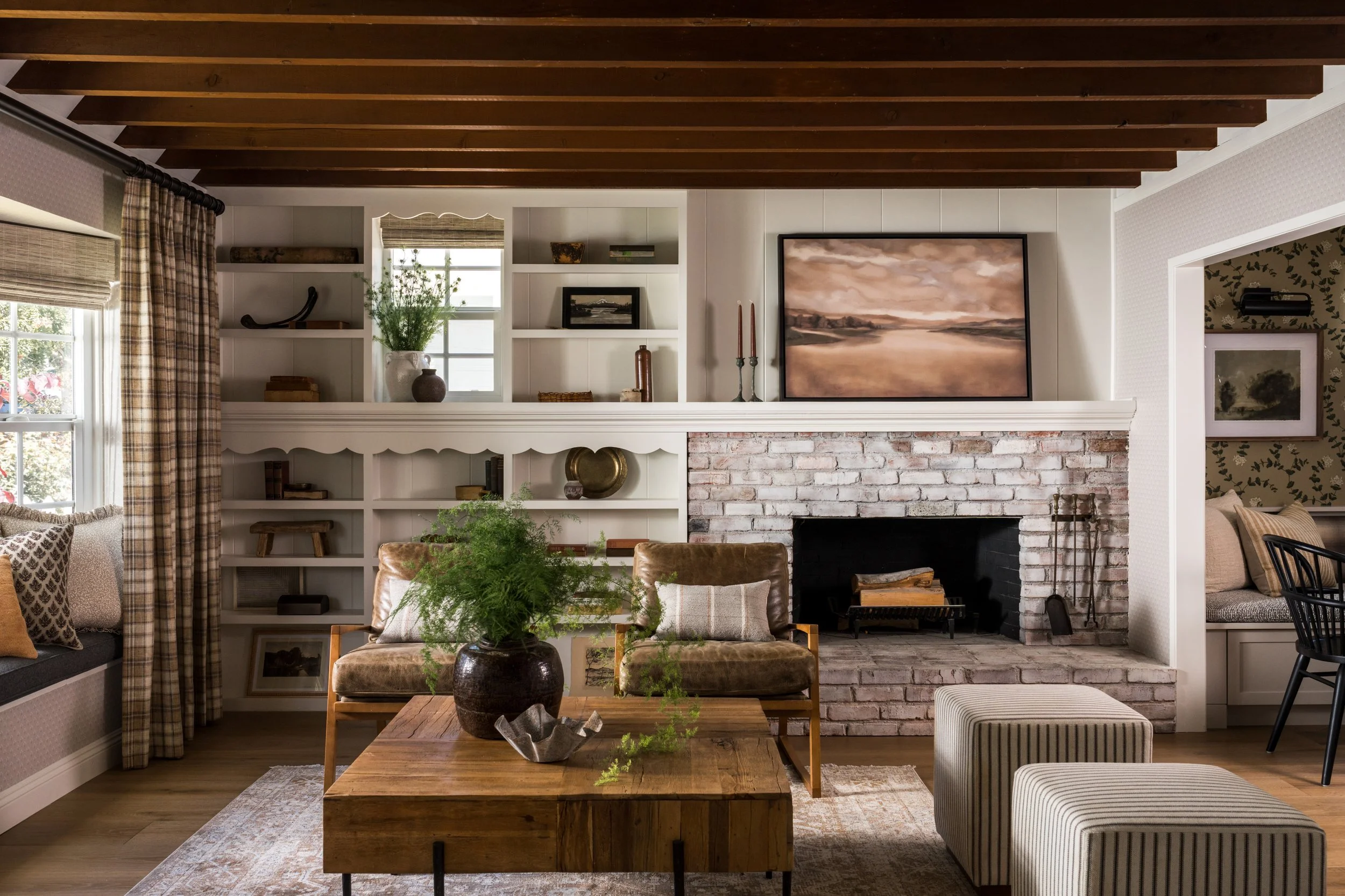 Living room with white brick fireplace, built-in white shelves, wooden ceiling beams, and cozy seating including leather chairs and ottomans.