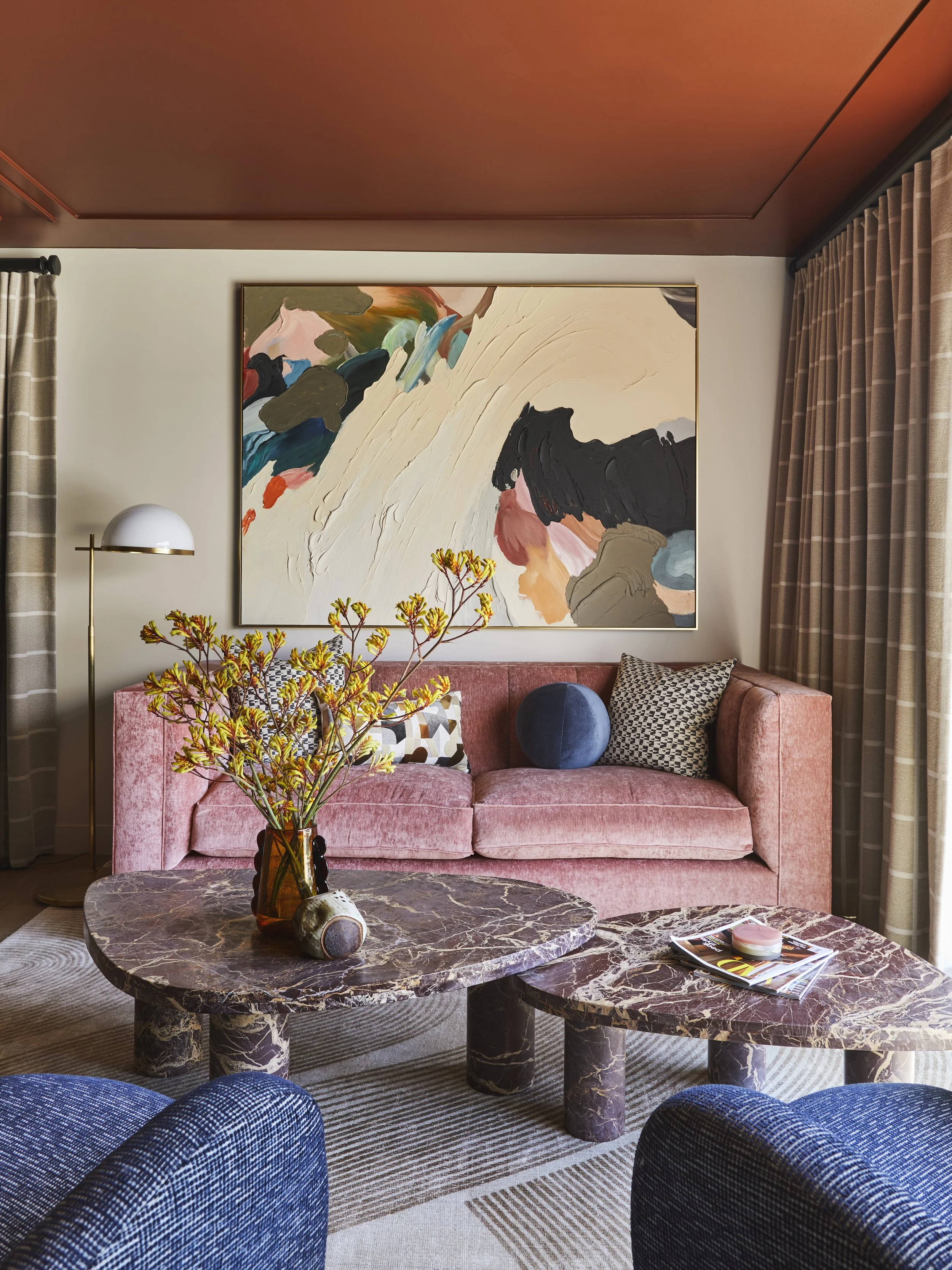 Living room with pink velvet sofa, abstract wall art, marble coffee tables, blue armchair, and beige curtains.