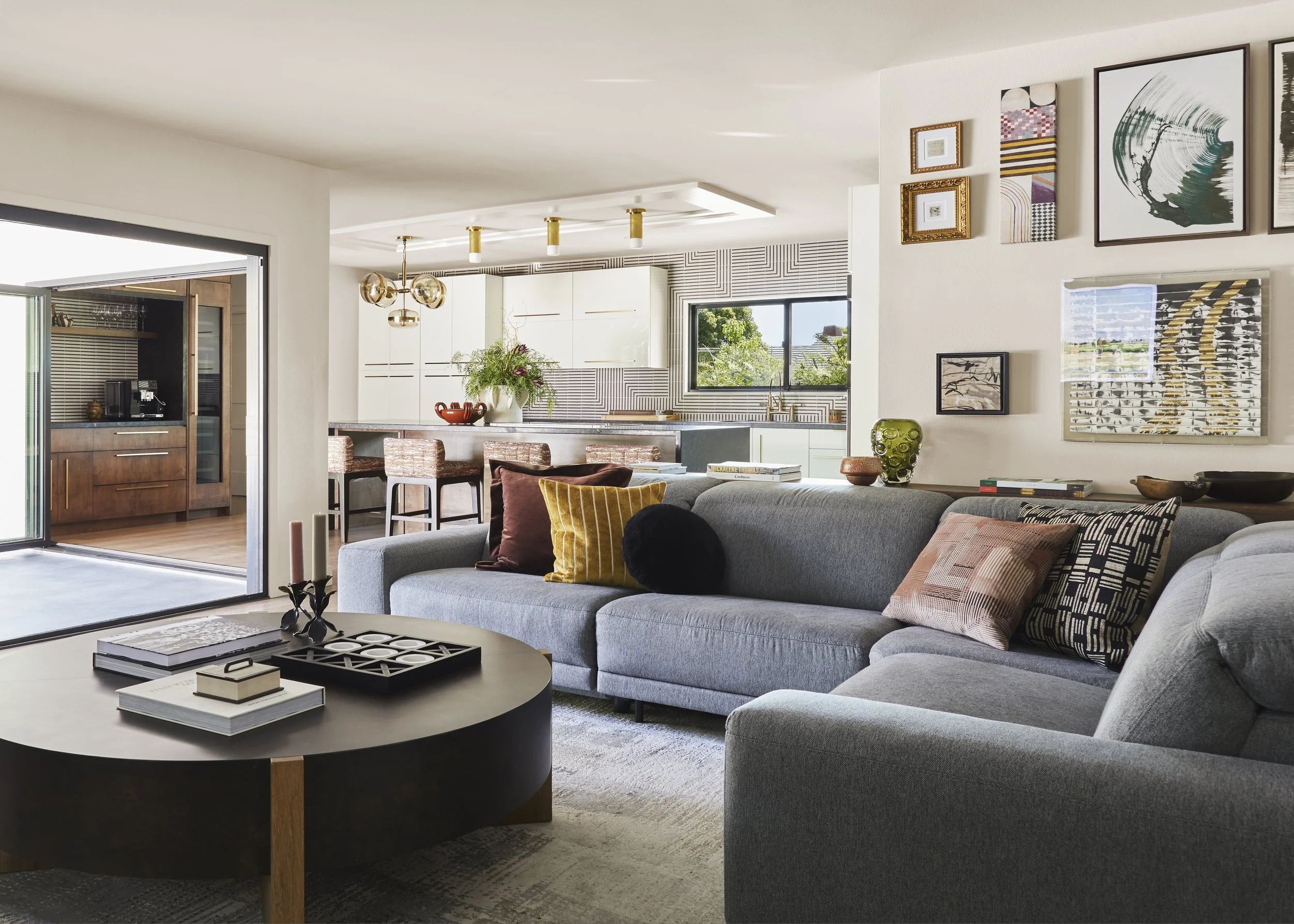Living room with gray sectional sofa decorated with patterned and chromatic pillows, oval black coffee table with books and accessories, various art pieces on the wall, open kitchen in the background with seating, window view of a clear sky and trees.