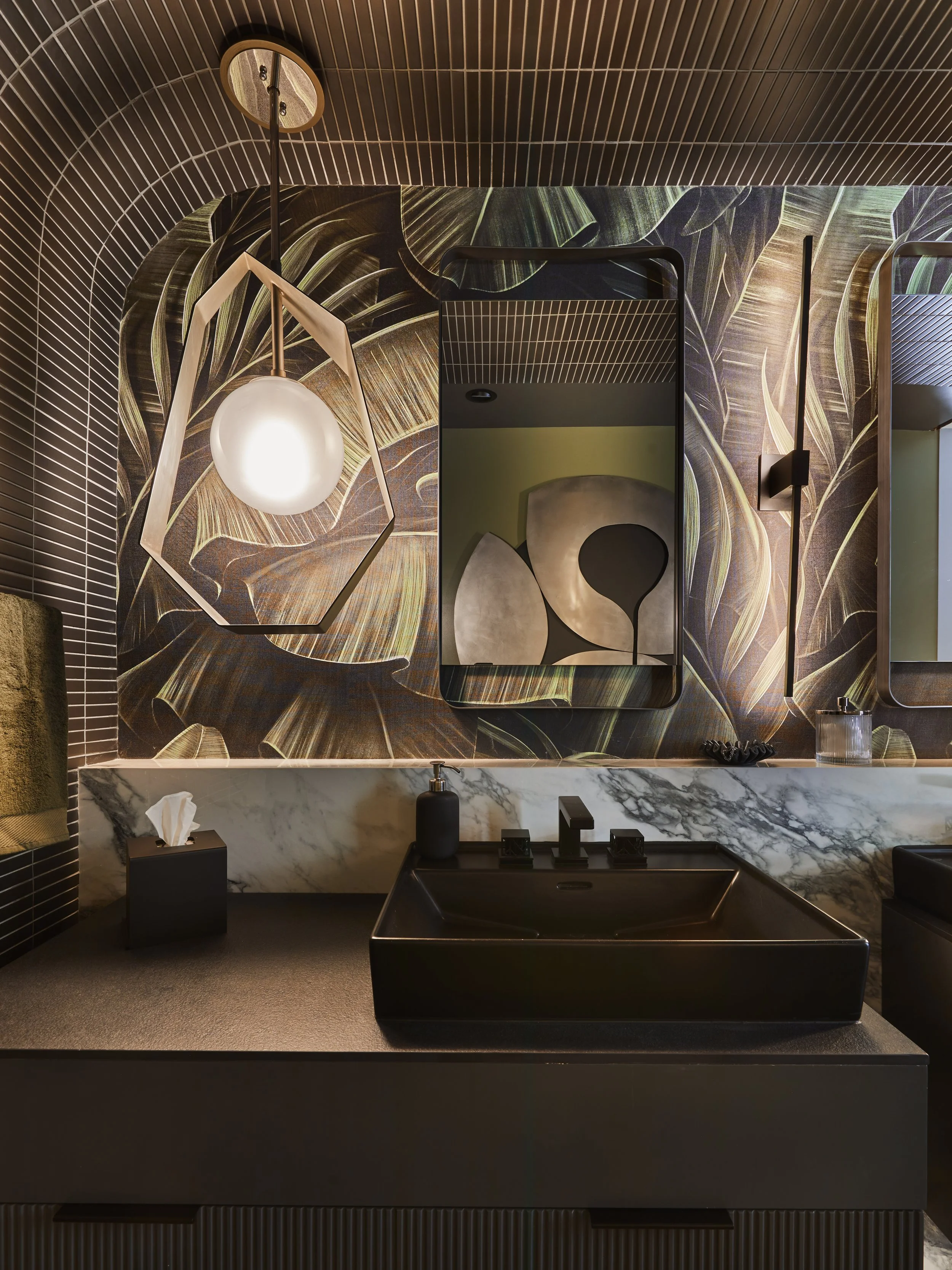 Modern bathroom vanity with a black sink, a black soap dispenser, and a tissue box on a marble countertop. Behind the countertop is a decorative wall with botanical patterns, a mirror, and modern lighting fixtures.