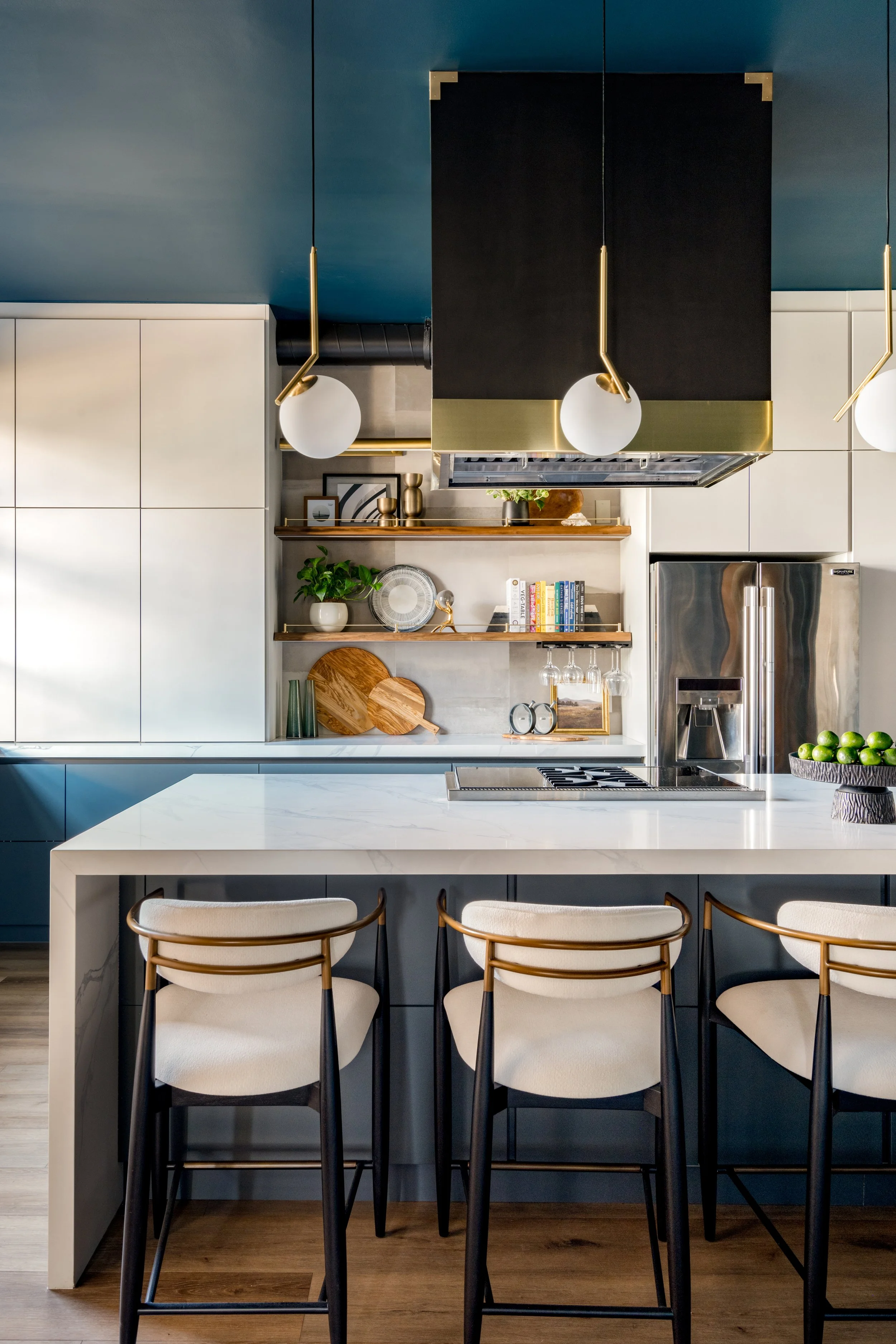 Modern kitchen with blue and white cabinetry, open shelves with decorative items, a stainless steel refrigerator, a white marble island with seating, and contemporary pendant lights.
