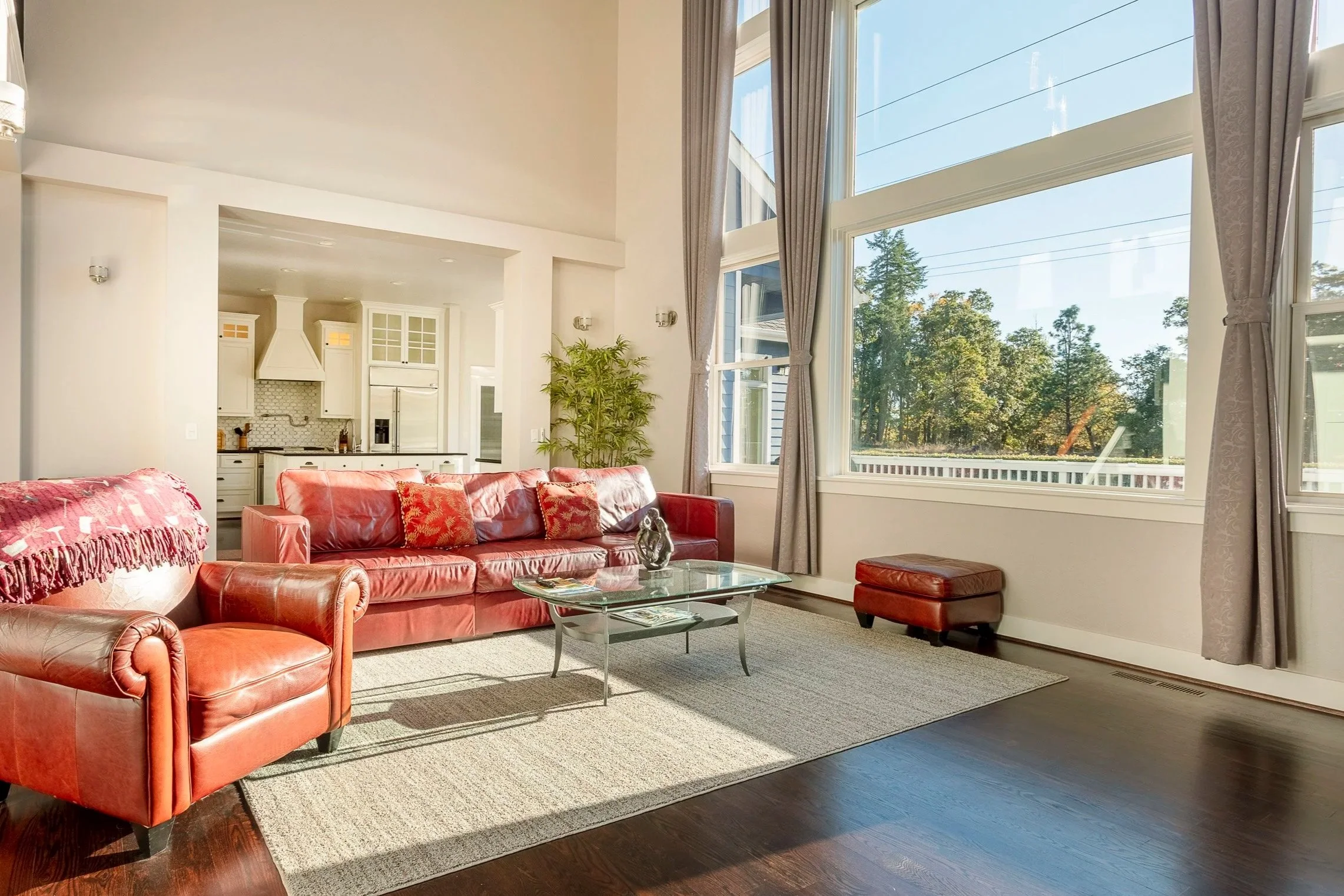 Living room with red leather sofa and armchair, glass coffee table, large windows with curtains, hardwood floors, view of trees outside, kitchen in background.