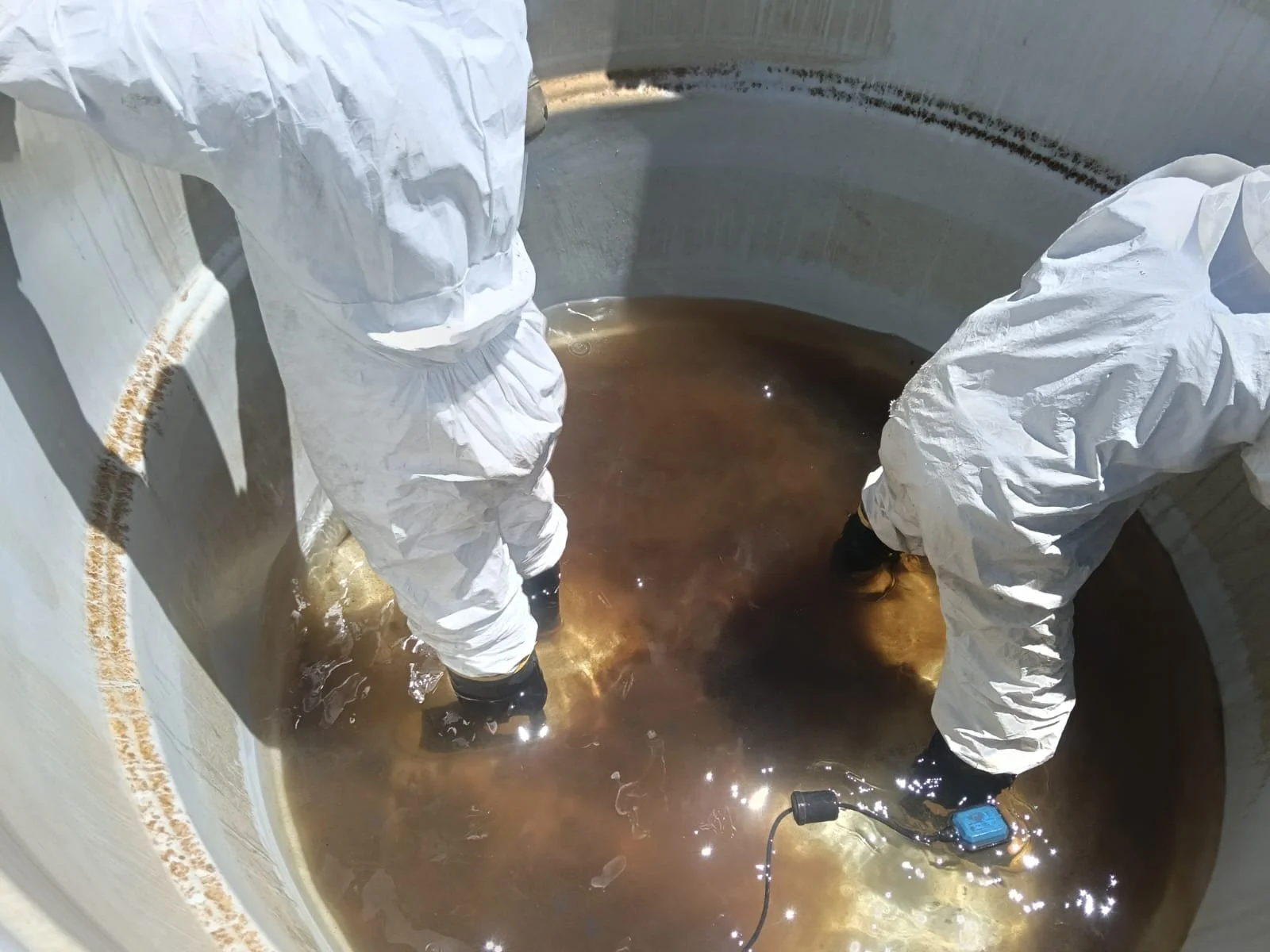 Dos personas con trajes de protección trabajan en un tanque grande con agua marrón, utilizando equipos especializados.