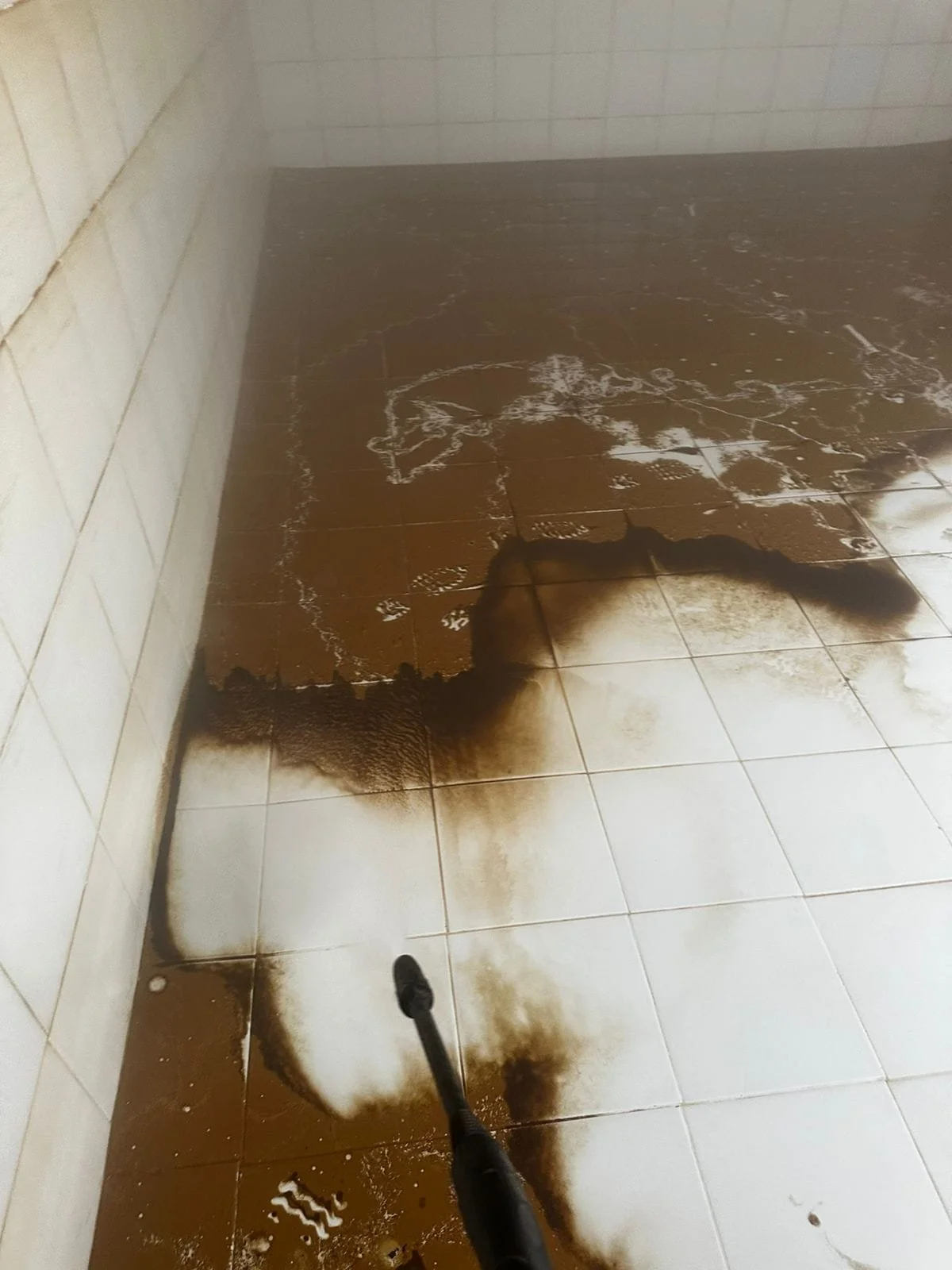 Piso de baldosas blancas y marrones en un baño, con manchas de humo negro y una manguera de presión que limpia el suelo dentro de un tanque de auga