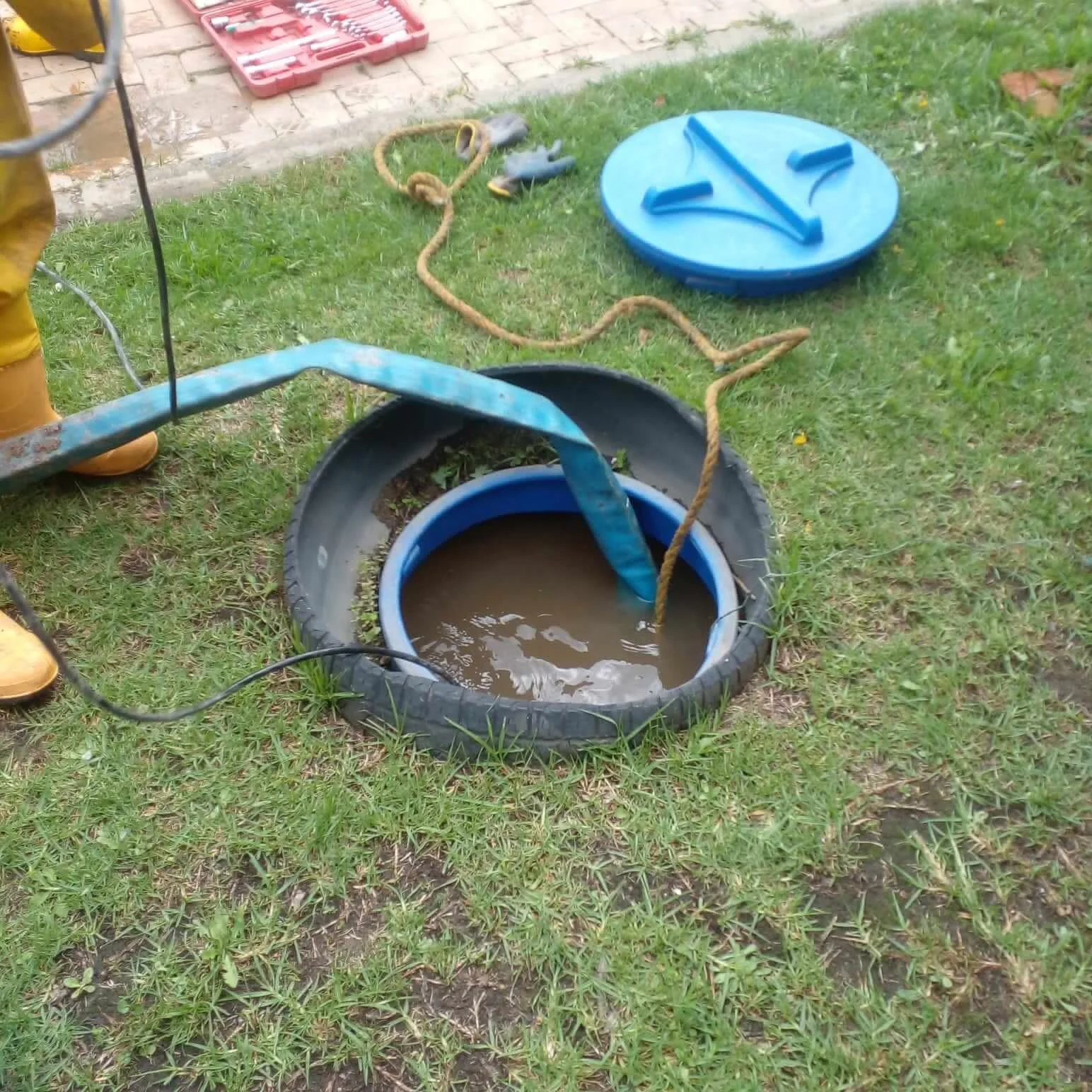 Tanque con agua sucia, manguera y tapa de plástico azul en un césped, indicando trabajo de limpieza o mantenimiento en un área exterior.