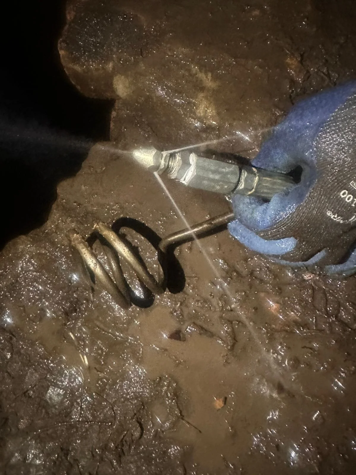 Persona usando guantes trabaja con herramienta en suelo fangoso y húmedo.