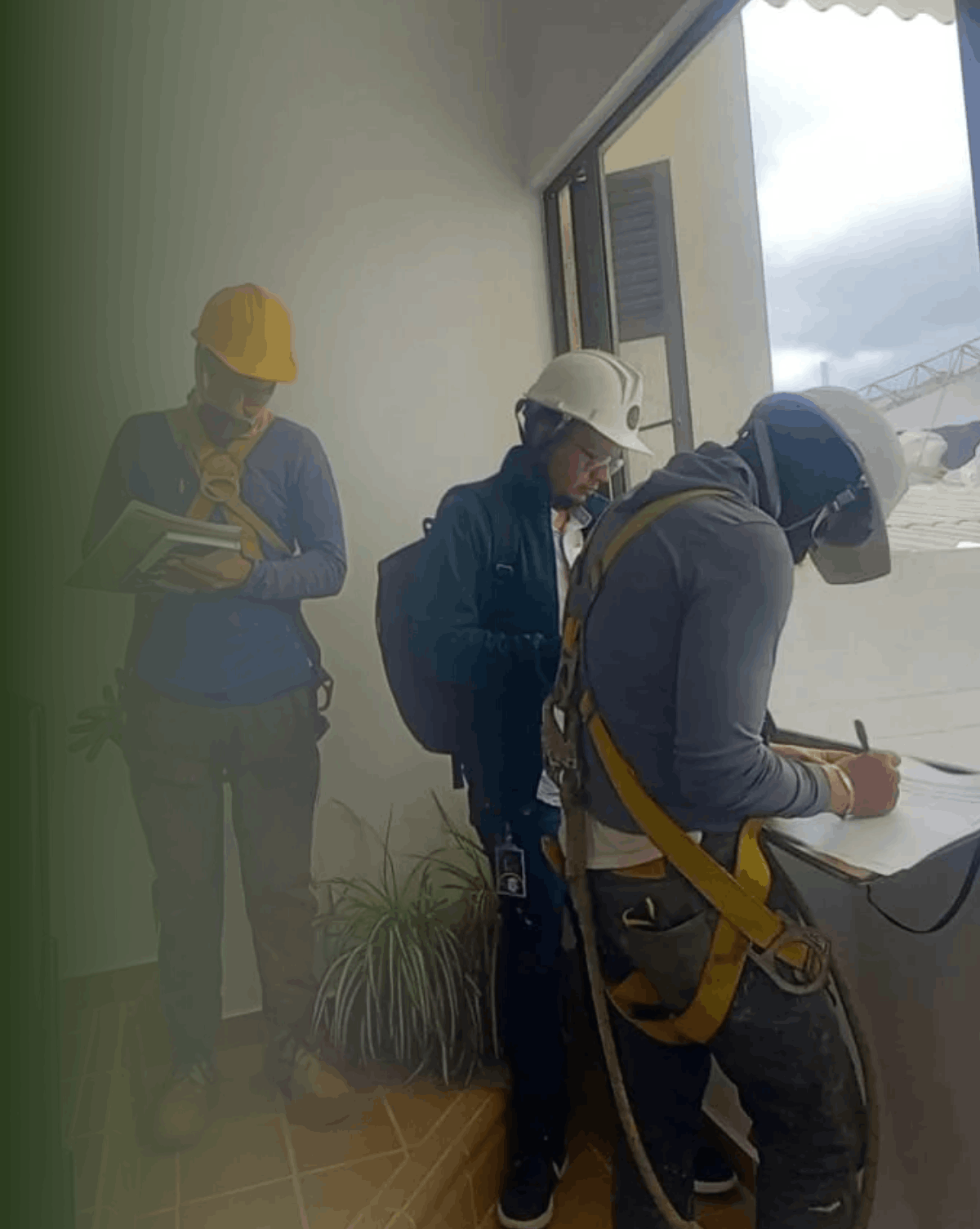 Tres trabajadores con cascos de seguridad y arnés, uno escribiendo en un papel, en una oficina con una ventana y plantas, rodeados de polvo y escombros.