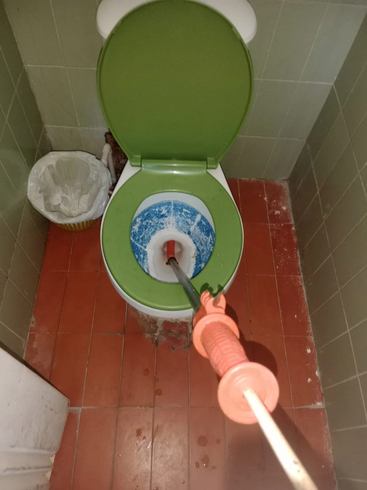 Agua siendo vertida en un inodoro con asiento verde en un baño con piso de azulejos rojos y paredes de azulejos verdes, con un bote de basura a la izquierda.